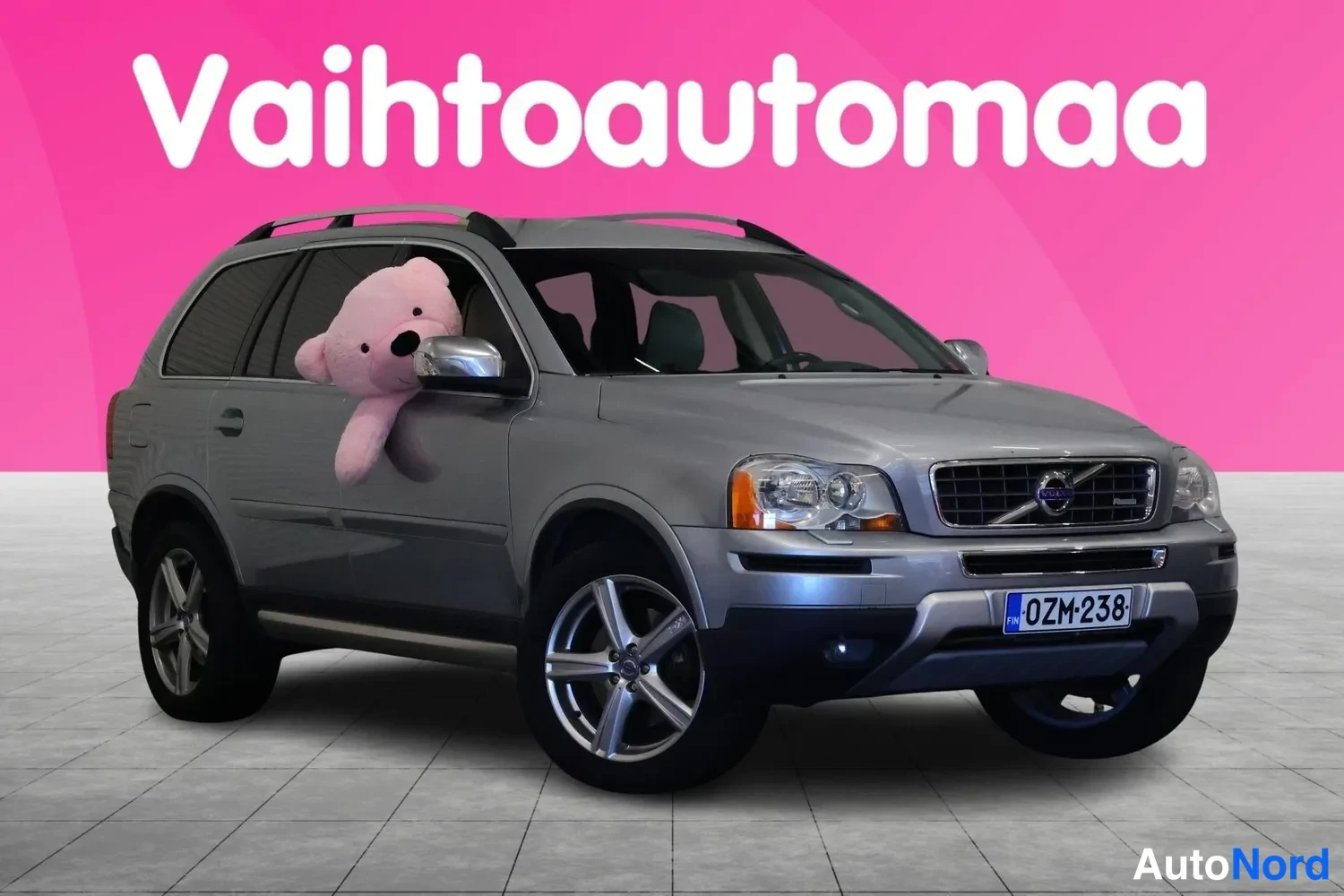 Etuviisto, nokka oikealle: Hopeanharmaa Volvo XC90 etuviistöstä oikealta, rekisteritunnus, kattokaiteet ja pinkki tausta.