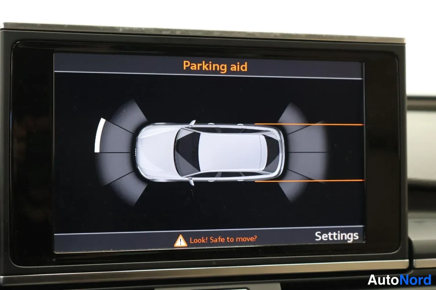 Yksityiskohta: Autoin päällä: MMI-näyttö, jossa Parking aid -näkömää sekä auton yläkuvan ympärillä tutkasektoreita. Ilmoitus 'Look! Safe to move?'. Kellonaika ei näy.