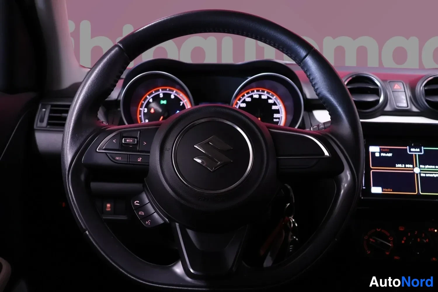 Muu: Ohjauspyörän tarkempi näkymä Suzuki-merkillä ja_MULTIpainikkeilla, taustalla osa mittaristosta ja infotainment-näytöstä.