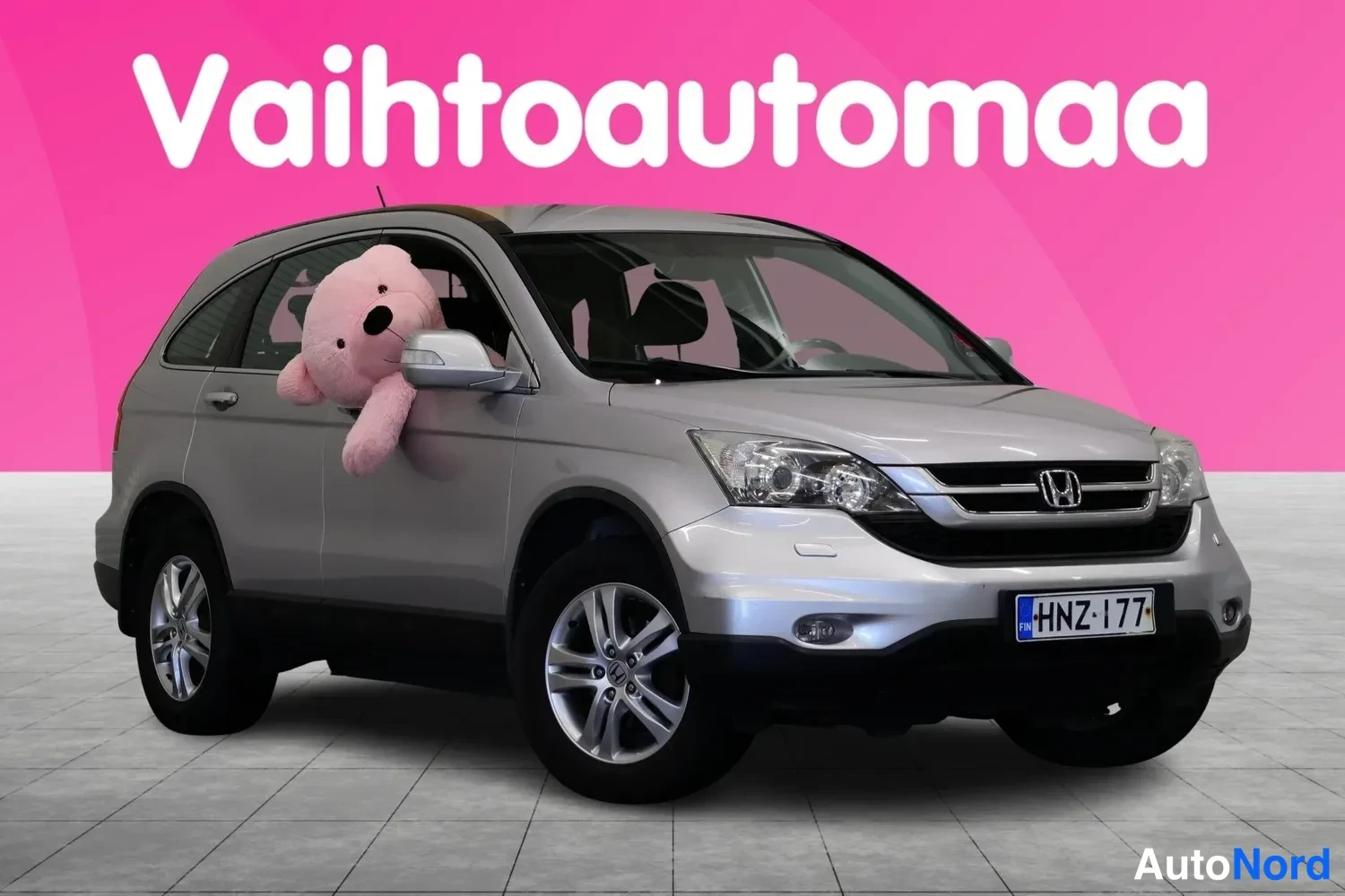 Etuviisto, nokka oikealle: Hopeanvärinen Honda CR-V etuosa kolmannesvasemmasta oikealta kuvattuna, rekisteritunnus, vaaleanpunainen tausta tekstillä Vaihtoautomaa.