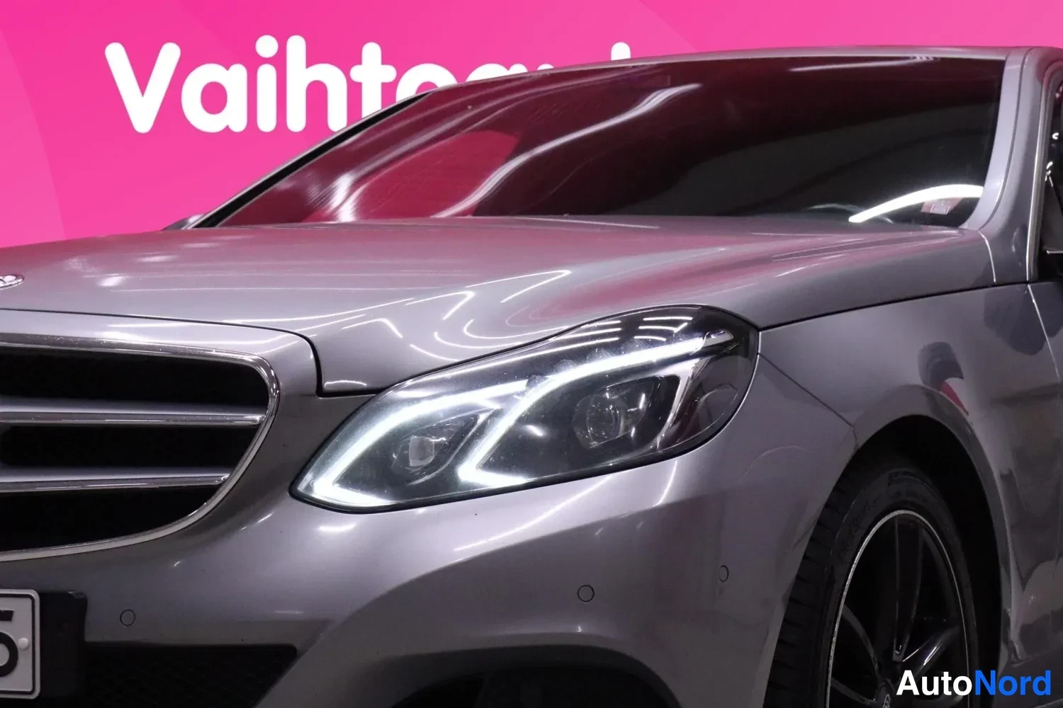 Edestä: Lähikuva Mercedes-Benzin etupäästä; ajovalo ja etusäleikkö näkyvät, korin väri hopeanharmaa.