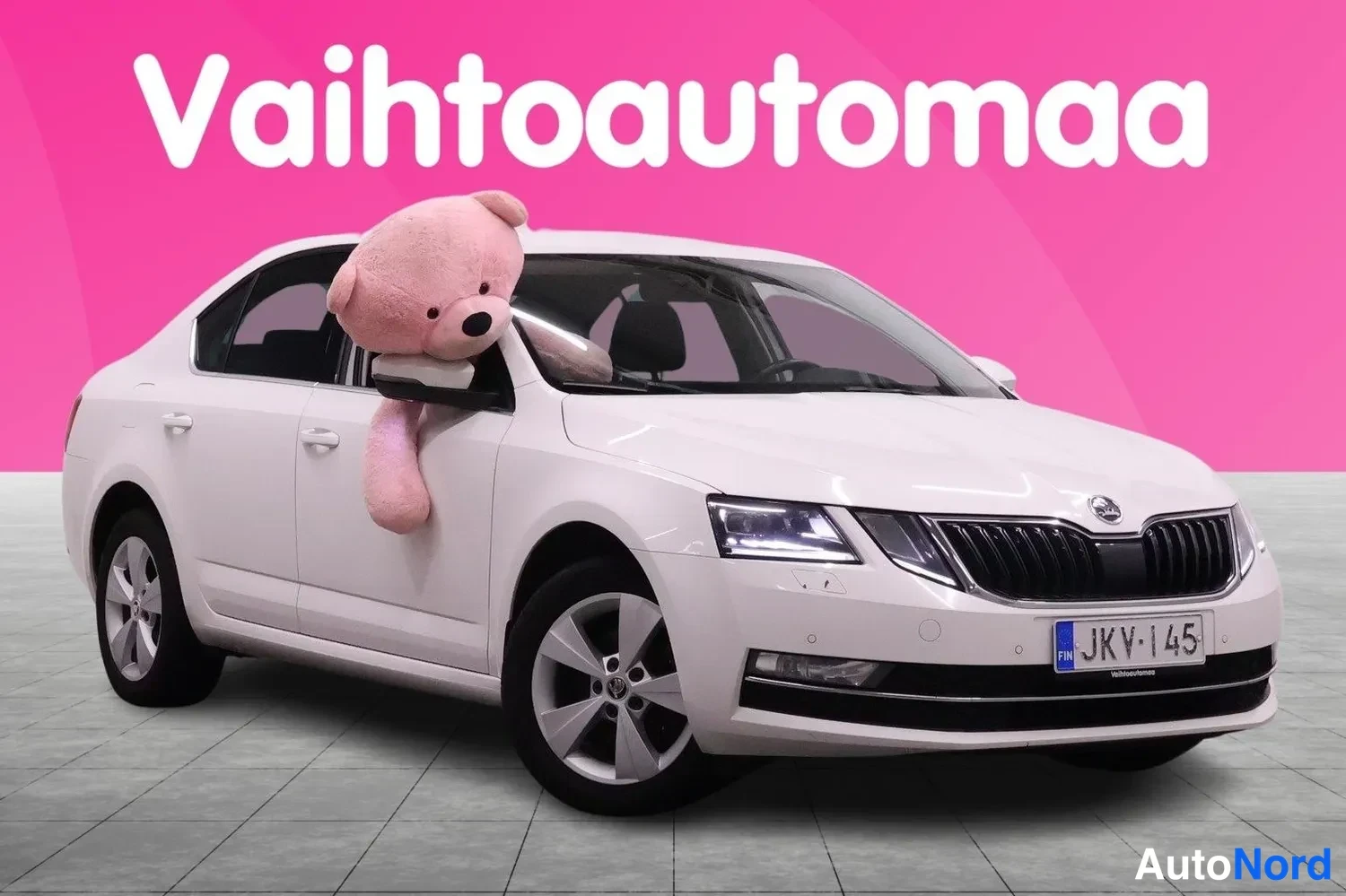 Etuviisto, nokka oikealle: Valkoinen henkilöauto kolmivartin edusta oikealta, rekisterikilpi, pehmolelu ikkunassa, vaaleanpinkki tausta.