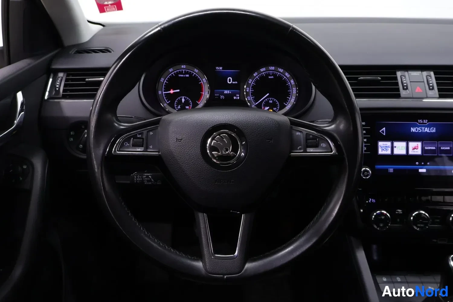 Muu: Lähikuva Skoda-ohjauspyörästä ja mittaristosta; keskitys näyttää 0 km/h ja kellonajan 15:32, oikealla osa kosketusnäytöstä tekstillä NOSTALGI.