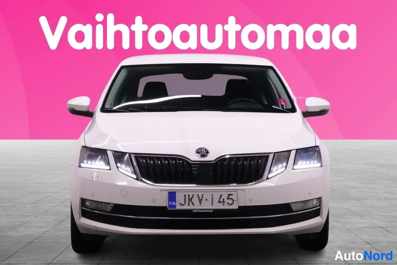 Edestä: Valkoinen henkilöauto suoraan edestäpäin, rekisterikilpi, vaaleanpinkki tausta.
