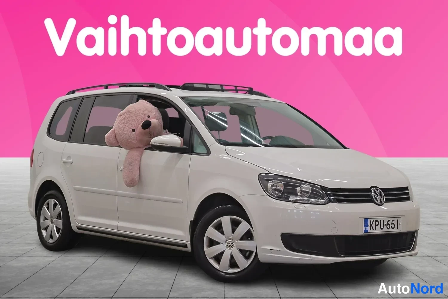Etuviisto, nokka oikealle: Valkoinen Volkswagen Touran kolmesta neljäsosasta edestä oikealta kuvattuna, kattokaiteet, rekisterikilpi, pehmolelu etuikkunassa.