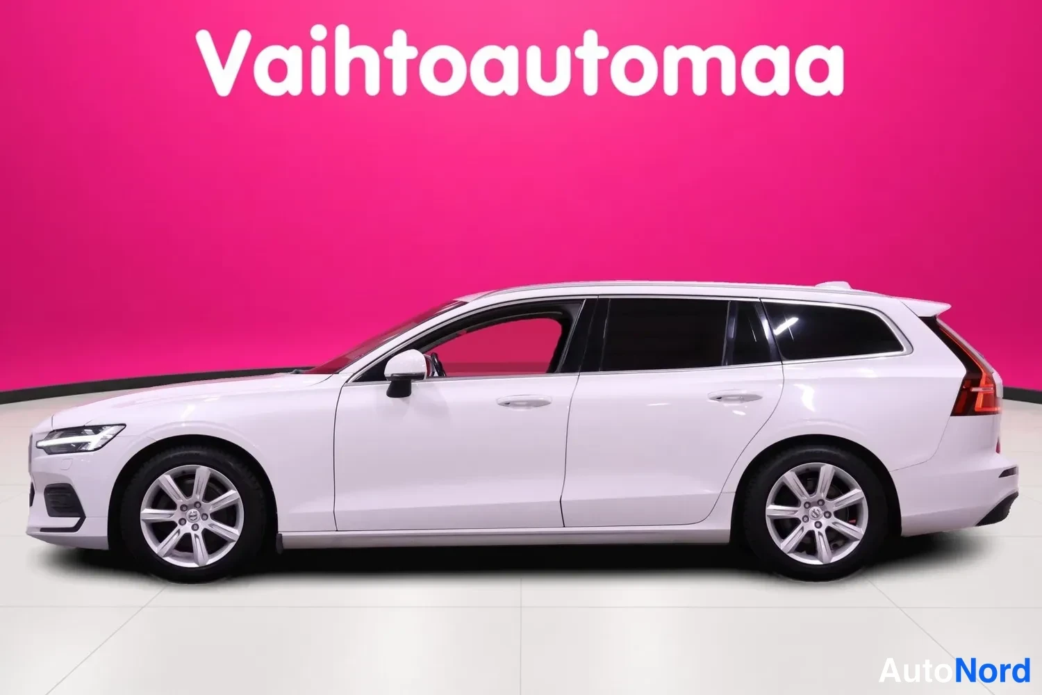 Etuviisto, nokka vasemmalle: Valkoinen farmariauto sivusta hieman etukulmasta, vasen kylki ja etummainen ajovalo näkyvissä, vaaleat vanteet.