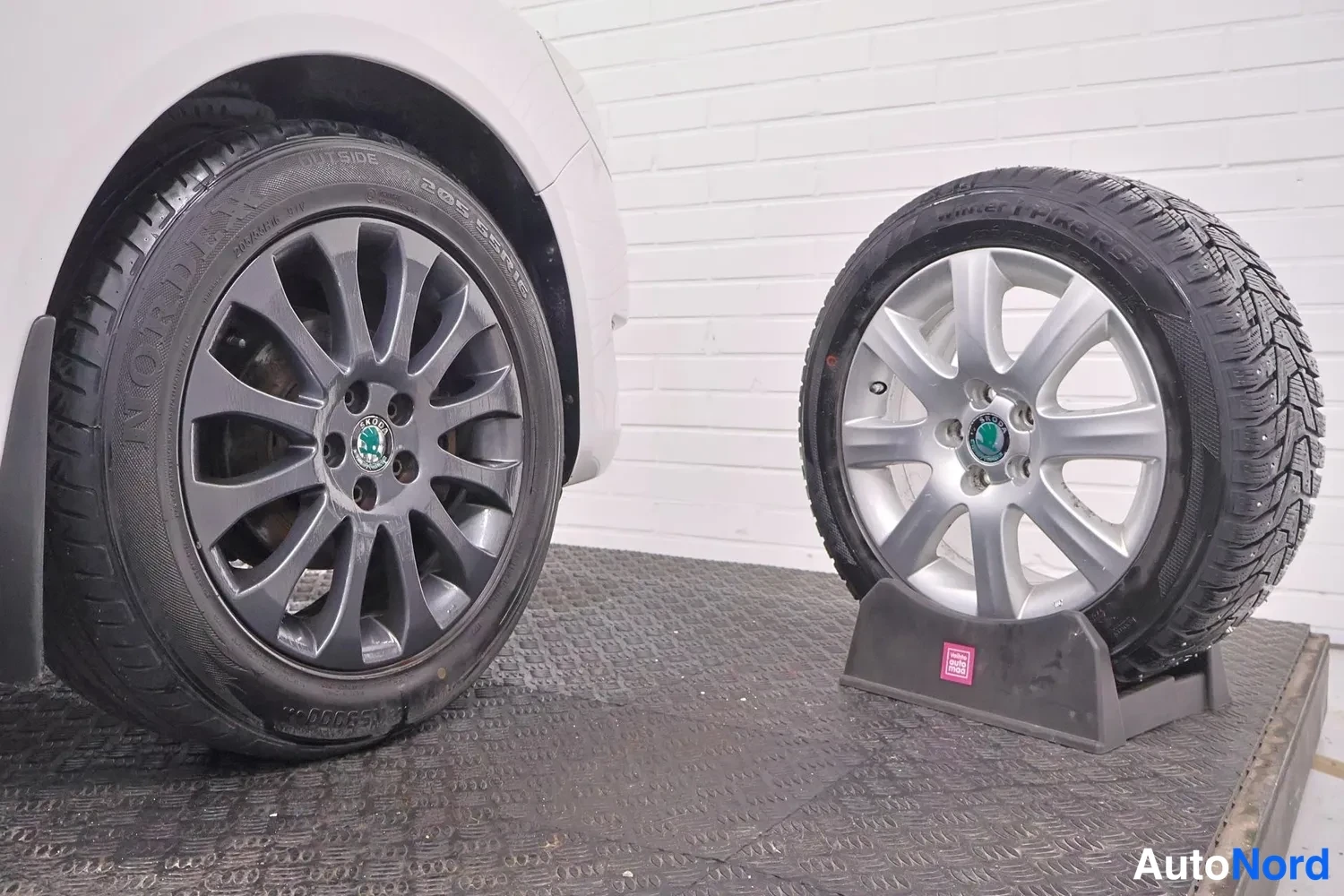 Muu: Auton tumma vanteella varustettu rengas ja vieressä jalustalla hopeavanteinen talvirengas merkityllä sidewall-tekstillä.