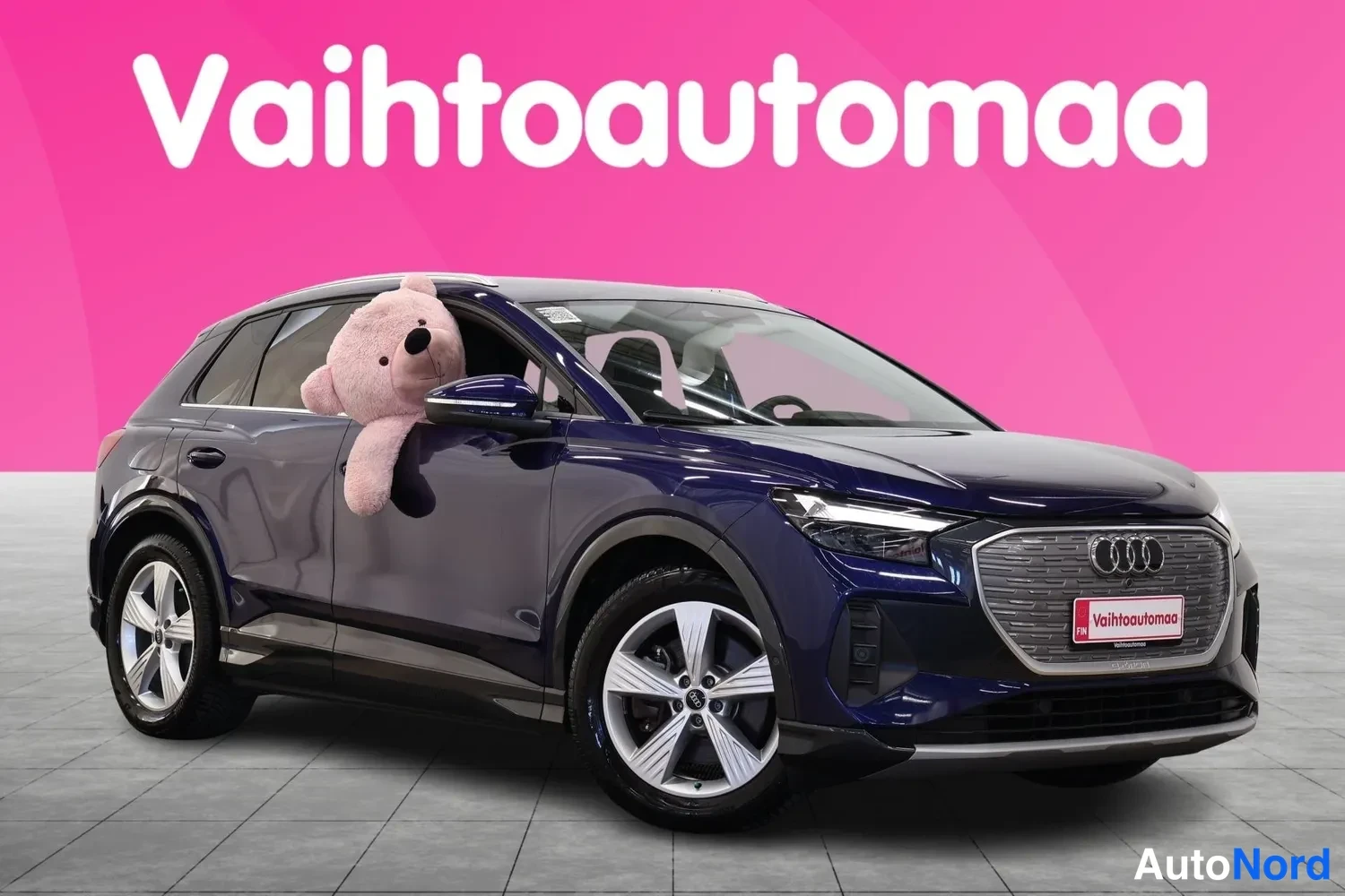 Etuviisto, nokka vasemmalle: Dark blue Audi SUV on a grey tiled showroom floor against a pink background. A light pink teddy bear is sitting in the open driver’s side window. The c