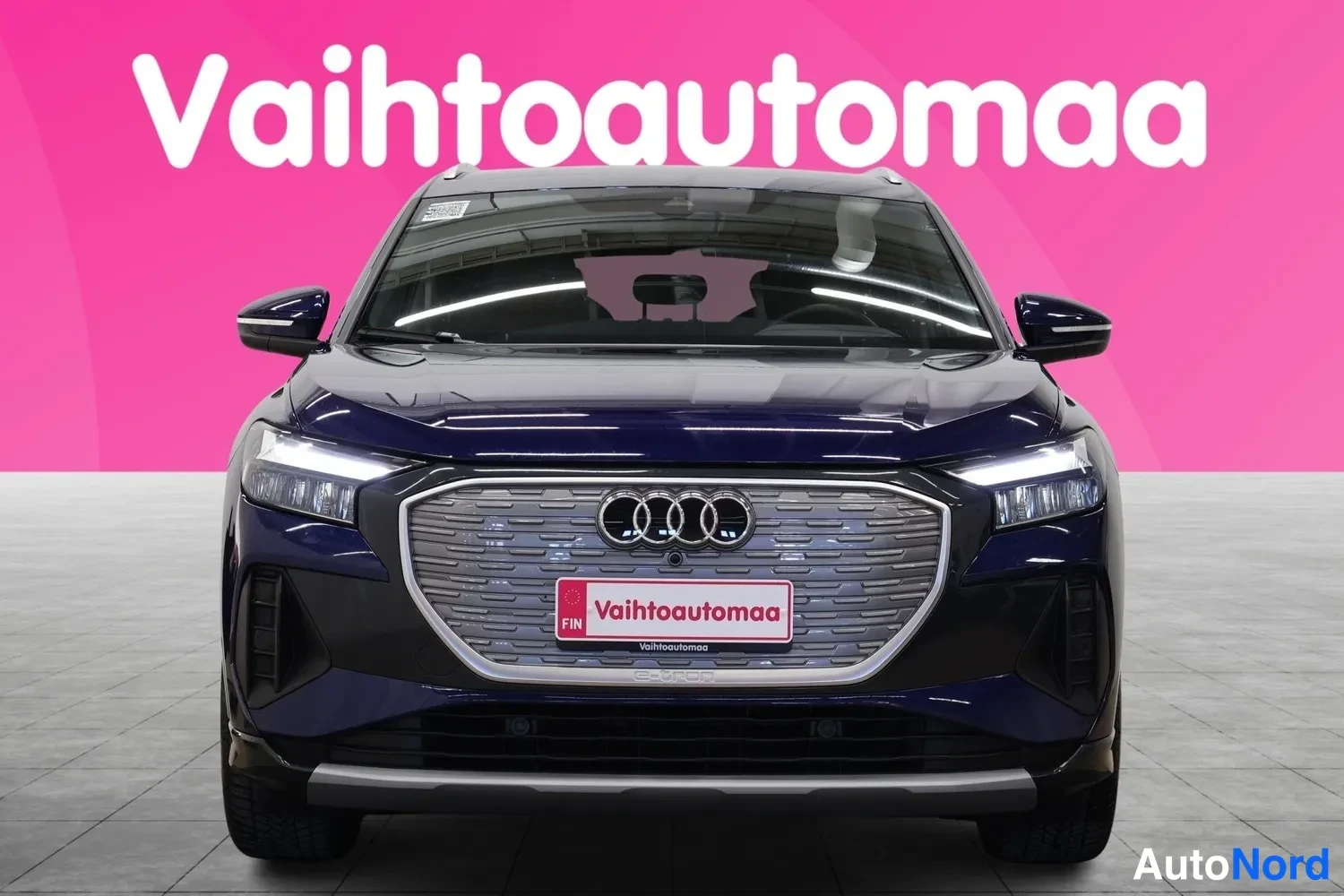 Edestä: Front view of a dark blue Audi SUV facing the camera directly against a pink studio wall. The headlights are illuminated and the four-ring Audi logo is centered