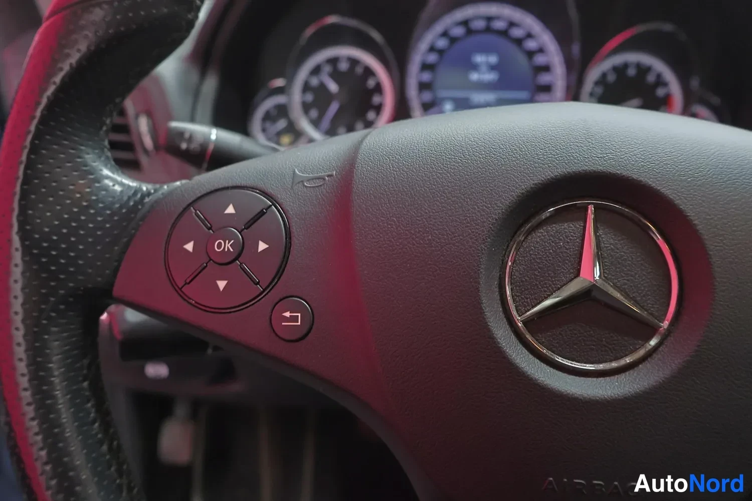 Yksityiskohta: Mustan nahkapintaisen Mercedes-Benz-ohjauspyörän vasen puolisko, jossa painikkeet: ohjausnuolet, OK-painike ja palautuspainike. Keskellä kolmionkulma-logo. Osia