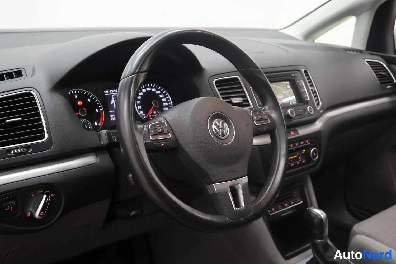 Muu: Volkswagenin ohjaamo: musta VW-ohjauspyörä, analogiset mittarit, multimedialaite näytöllä ja automaattivaihteiston valitsin.
