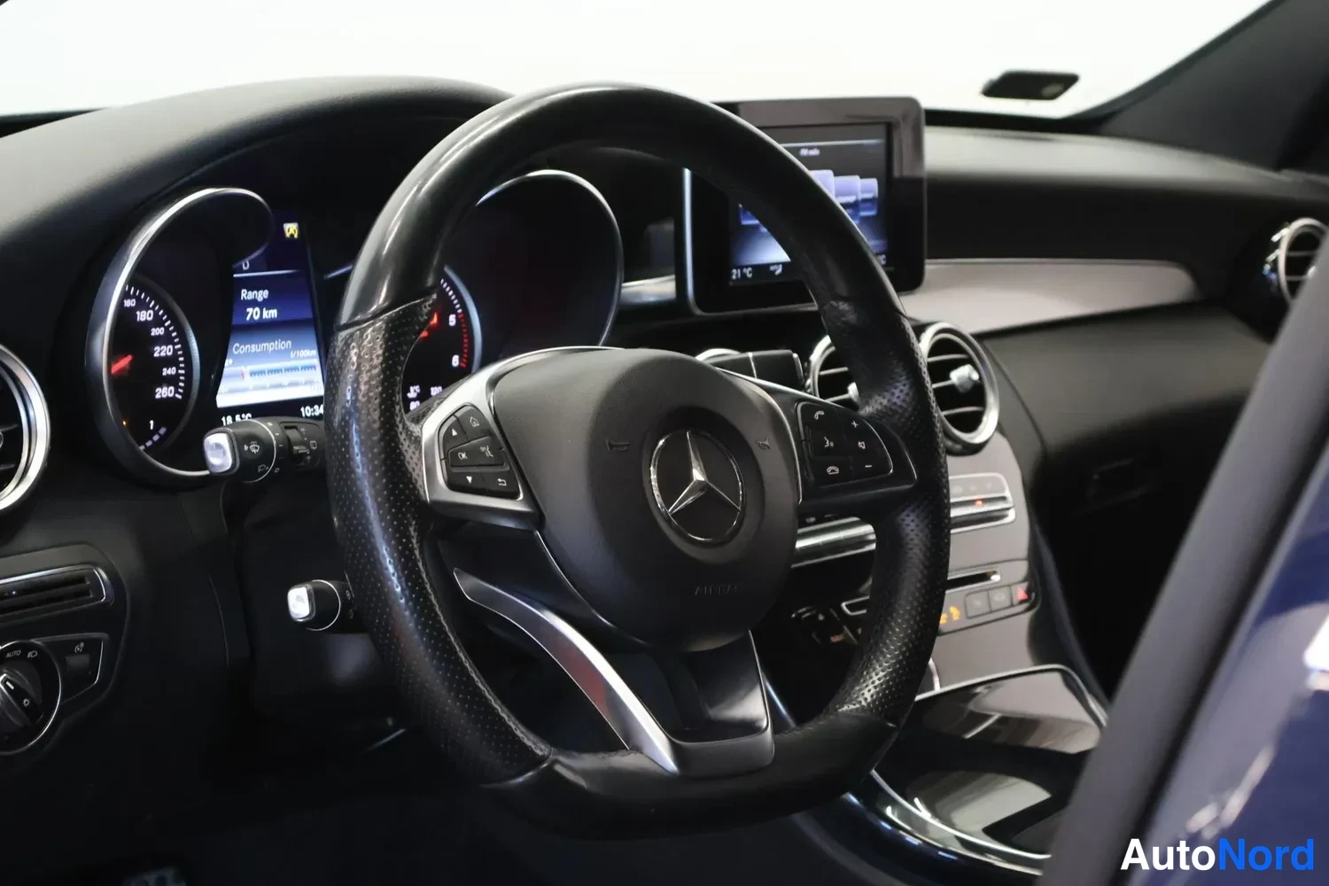 Edestä: Ohjaamon näkymä: musta Mercedes-merkillä varustettu ohjauspyörä, mittaristo, ilmastointisuuttimet ja keskusnäyttö.