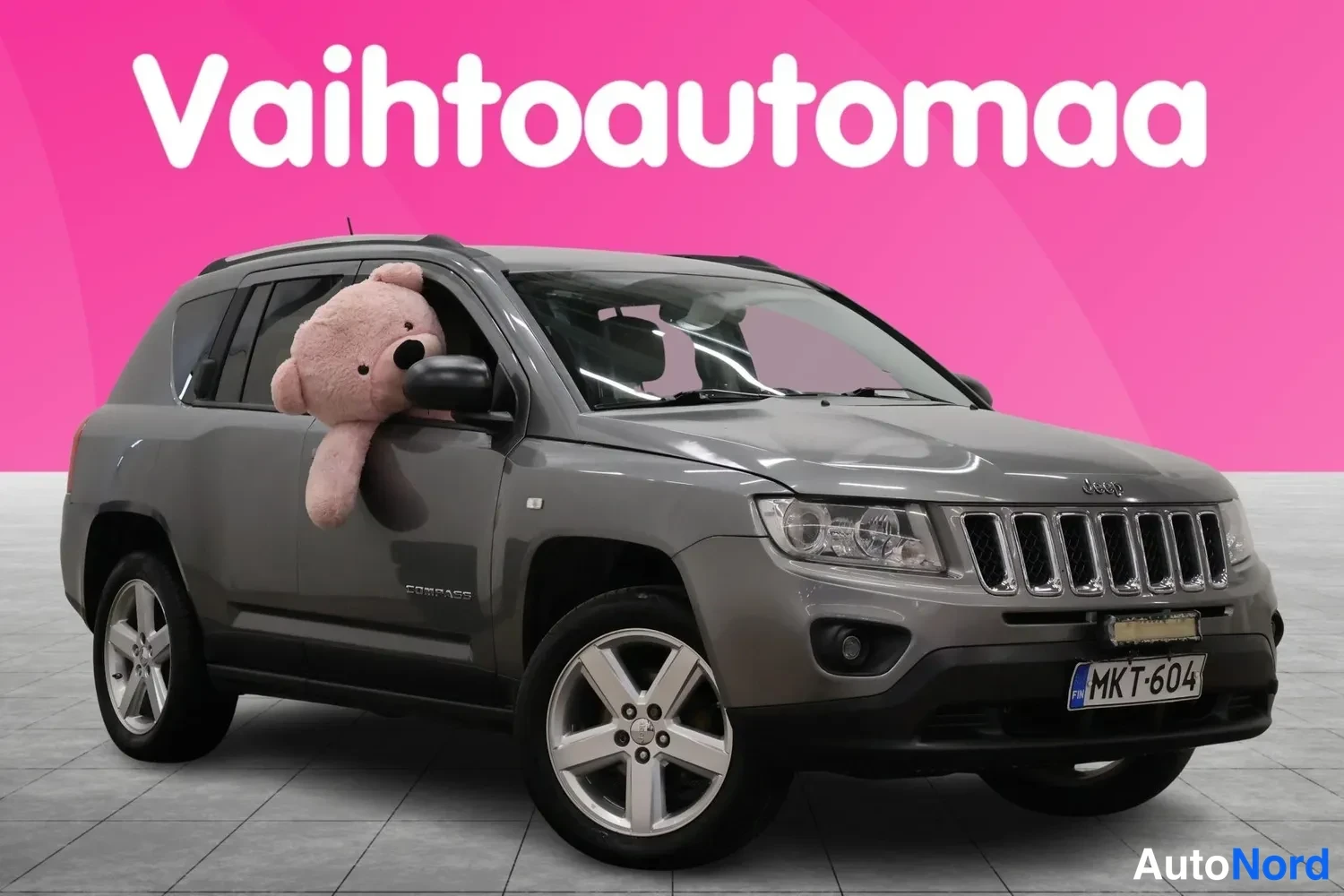 Etuviisto, nokka oikealle: Harmaa Jeep Compass etuviistöstä oikealle, rekisteritunnus, vaaleanpunaisessa studiotaustassa.
