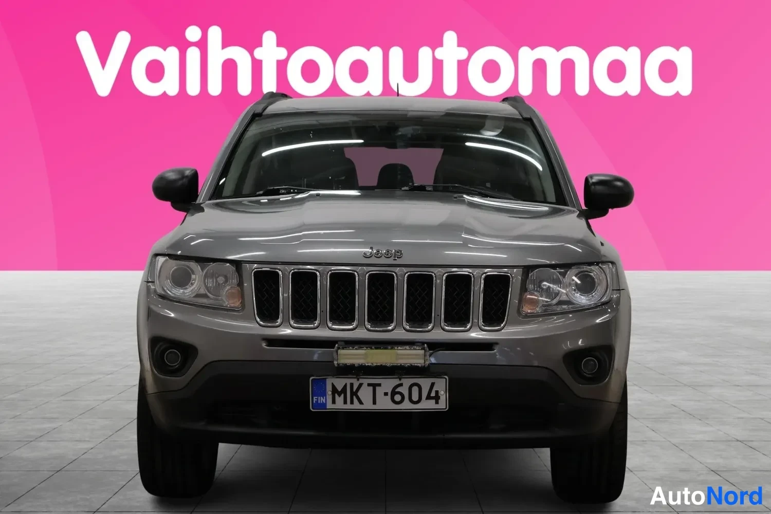 Edestä: Harmaa Jeep Compass suoraan edestä, rekisteritunnus, vaaleanpunaisessa studiotaustassa.
