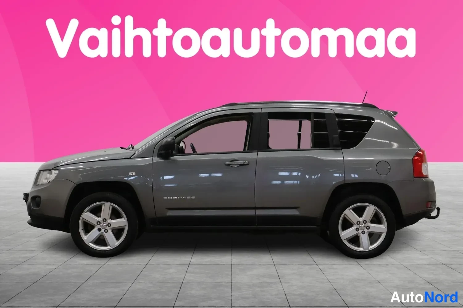Etuviisto, nokka vasemmalle: Auton etukulma vasemmalta, tummanharmaa Jeep Compass, kytketty perävaunukoukku etupuskurissa.
