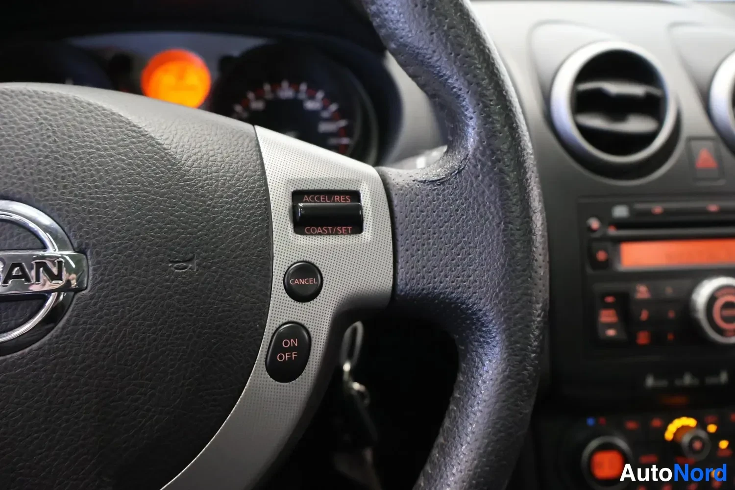 Muu: Nissan-ohjauspyörän lähikuva, jossa risteilyhallintanapit ACCEL/RES, COAST/SET, CANCEL ja ON OFF, taustalla kojelaudan ilmastointiaukot ja radiopaneeli.