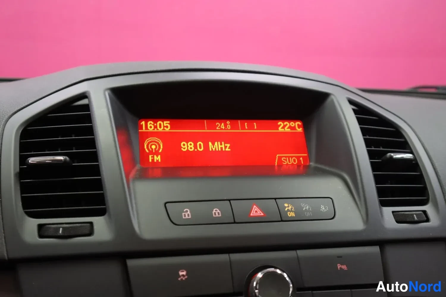 Muu: Kojelaudan näytön lähikuva: punainen FM-radionäyttö näyttää taajuutta 98,0 MHz, kelloa 16:05 ja lukemaa 22 °C; näytön alla varoituskolmio- ja lukkopainikkeet se