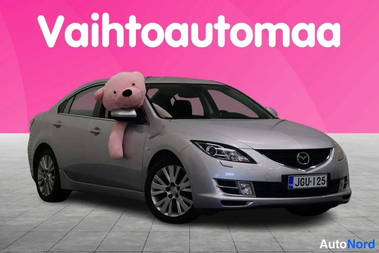 Etuviisto, nokka oikealle: Hopea Mazda 6 -henkilöauto kuvattuna etuviistosta, keula osoittaa kuvan oikealle reunalle. Etusäleikkö, ajovalot ja etupuskuri rekisterikilvellä näkyvät,