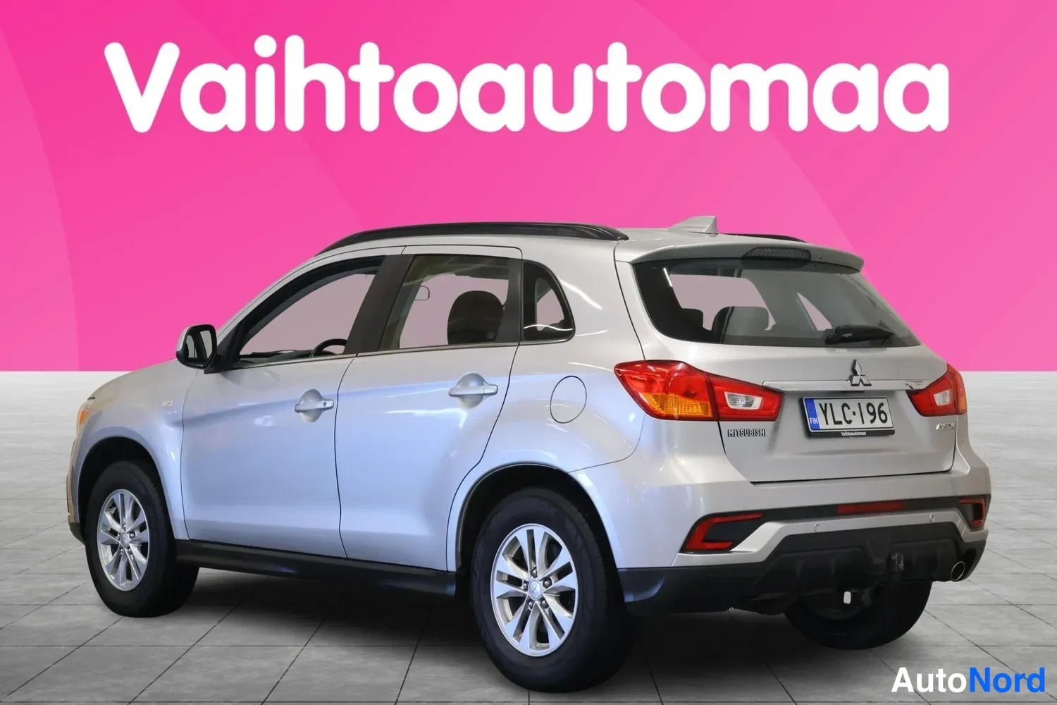 Takaviisto: Hopeinen hatchback taka­viistosta oikealta, rekisterikilpi, pinkki tausta.
