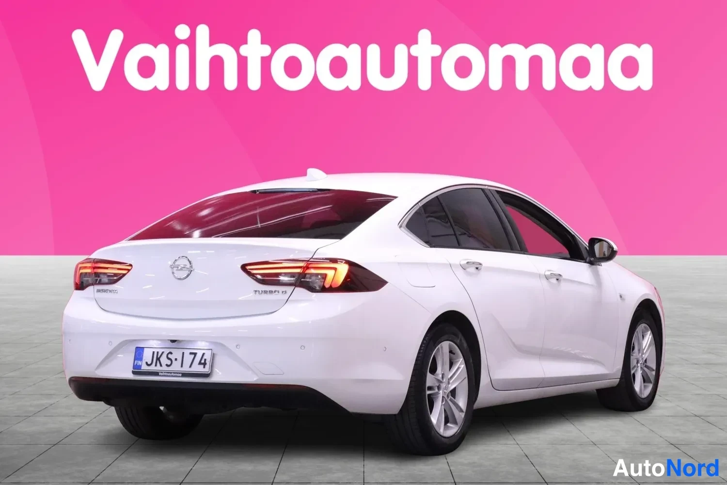 Takaviisto: Valkoisen Opel Insignian takaosa ja oikea kylki viistosti taakse; rekisteritunnus, taustalla vaaleanpunainen Vaihtoautomaa-mainos.