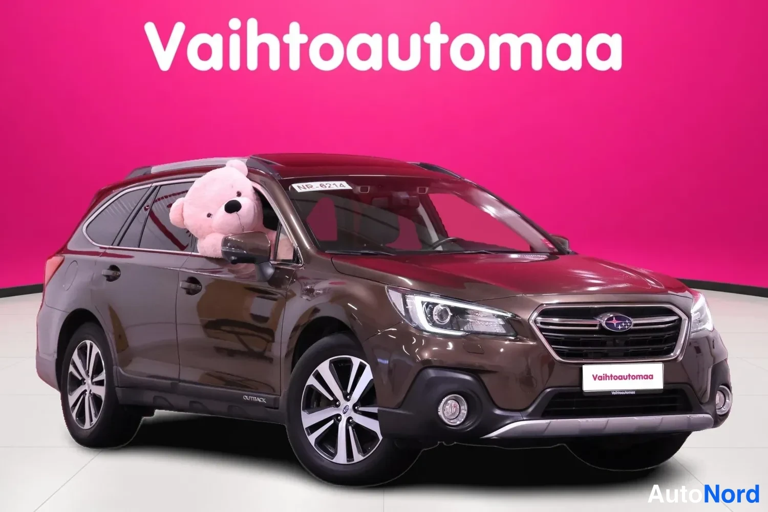 Etuviisto, nokka vasemmalle: Subaru Outback station wagon in metallic brown shown from a front three-quarter angle, with silver roof rails and alloy wheels visible against a pink s