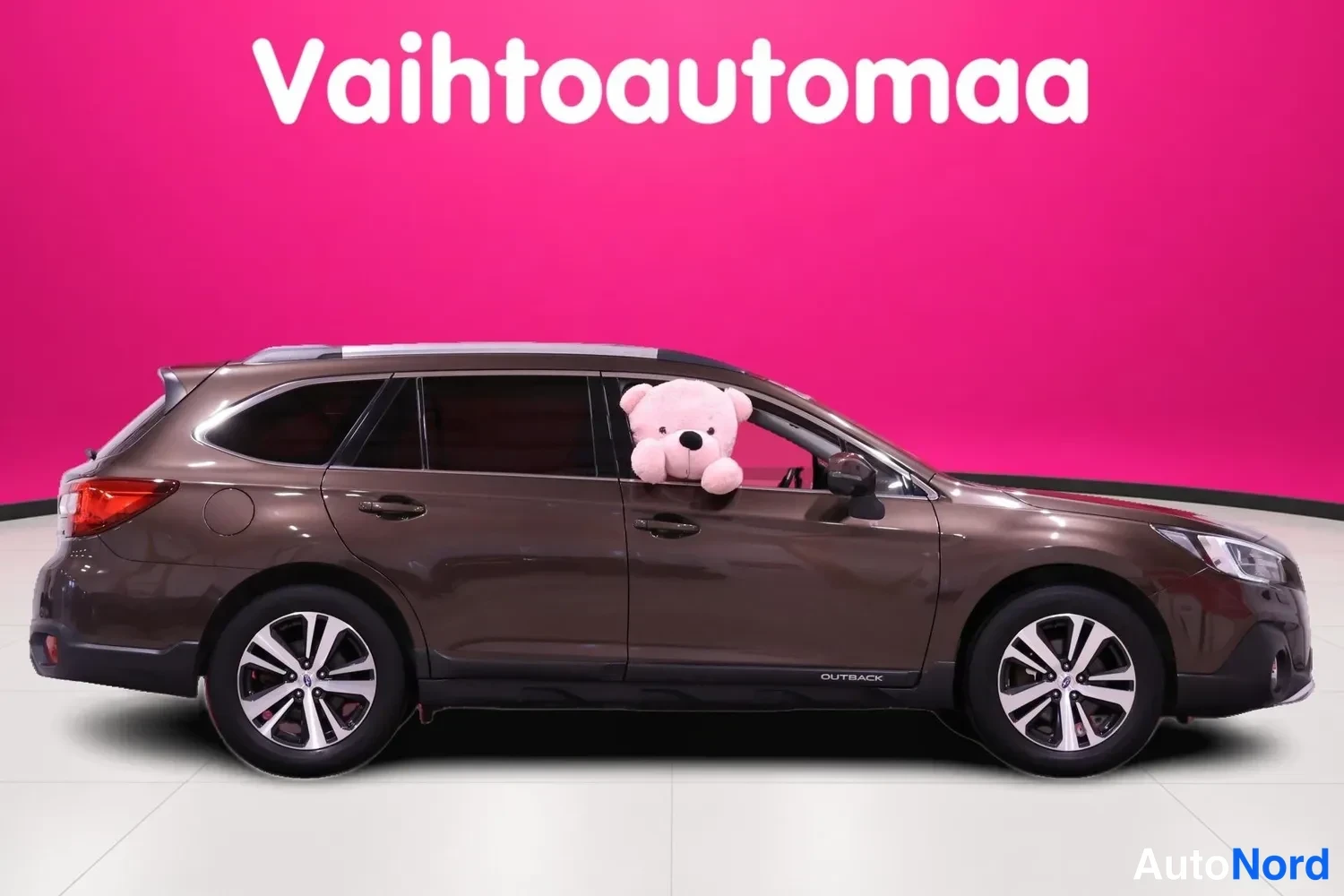 Sivulta: Side profile view of a Subaru Outback station wagon in metallic brown showing all four doors, tinted windows, and roof rails with a pink teddy bear in the drive