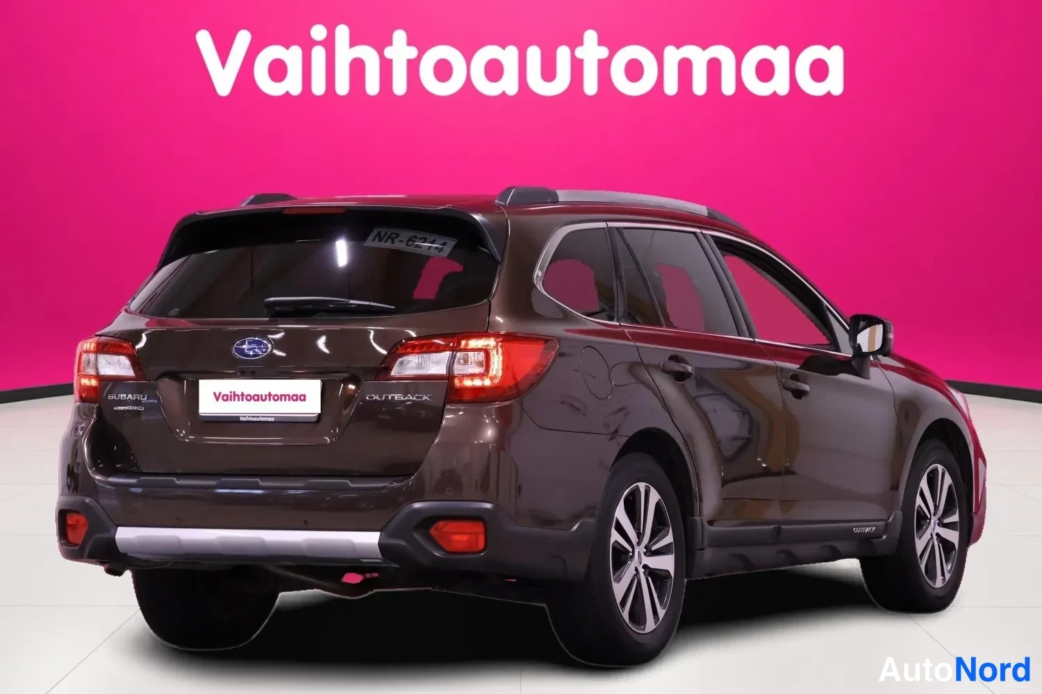 Takaviisto: Rear three-quarter view of a Subaru Outback station wagon in metallic brown displaying tail lights, rear bumper skid plate, and model badging on a pink backgrou