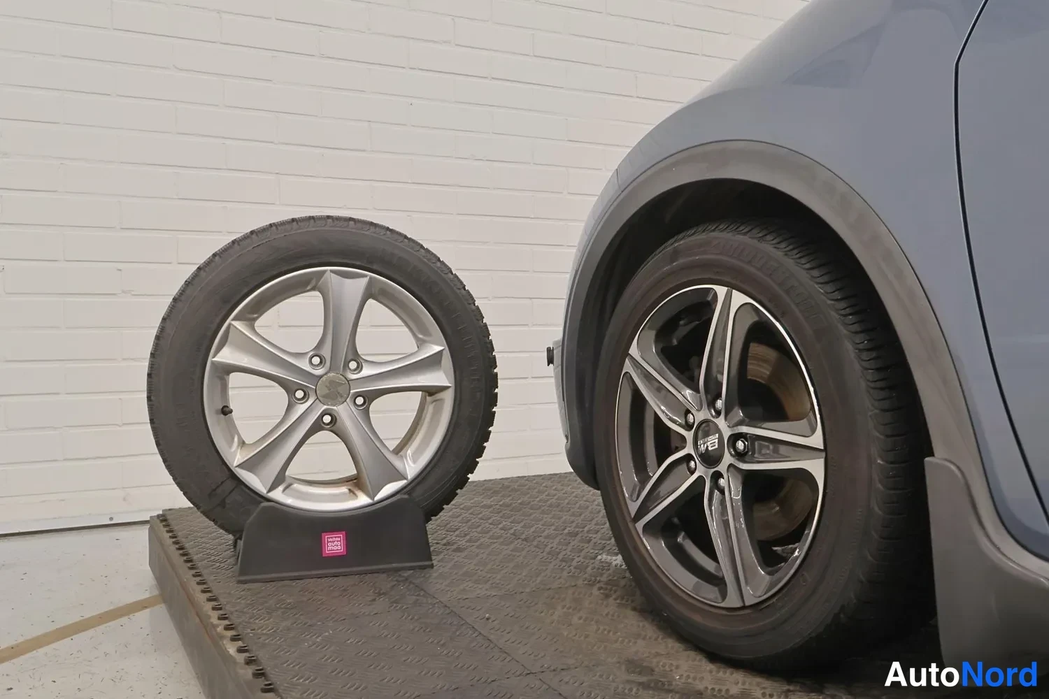 Yksityiskohta: Renkaanvaihtotilanne: japanilaistyylinen viisikanta-alumiinvanne rengas jalustalla ja autossa tumma viisikanta-vanne, harmaankylkinen auton etutakakulma.