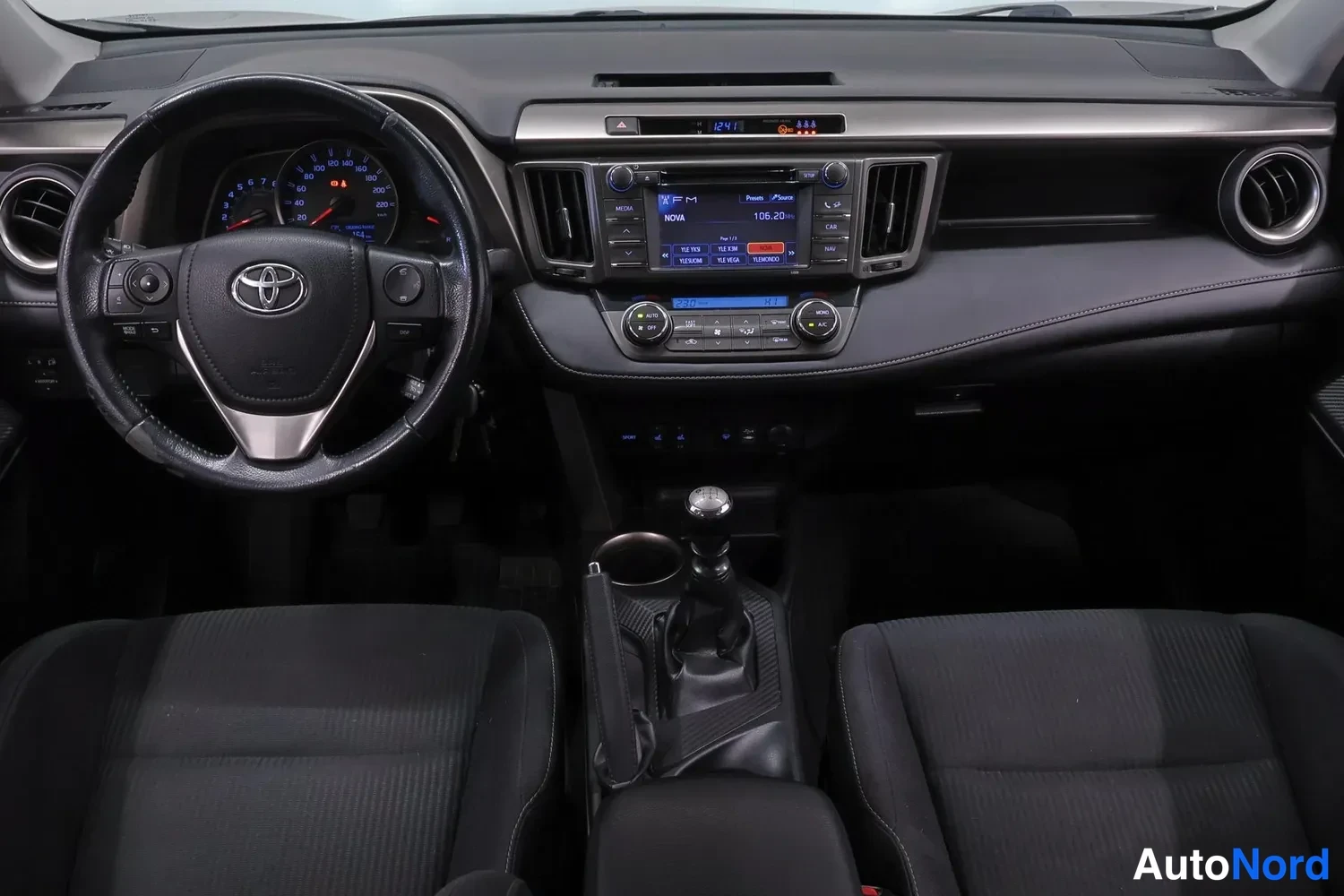 Muu: Kojelauta ja etuistuimet kuvattuna keskikäytävän yläpuolelta: Toyota-ohjauspyörä, analoginen mittaristo, kosketusnäytöllinen radio ja manuaalivaihteen keppi.