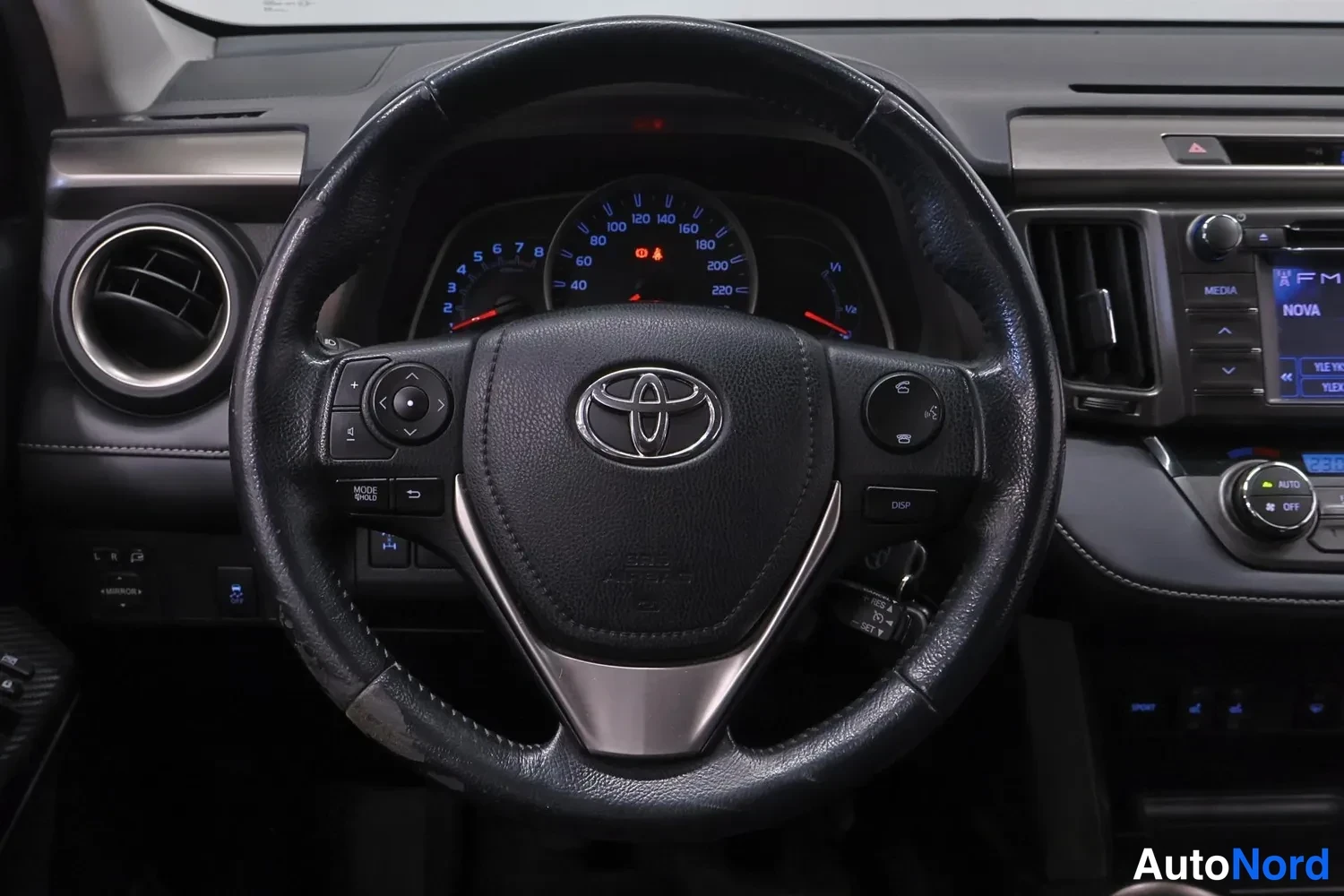 Muu: Ohjauspyörän ja kojelaudan läheiskuva: Toyota-logo ohjauspyörässä, mittaristo ja vasemmalla olevat nappiryhmät.