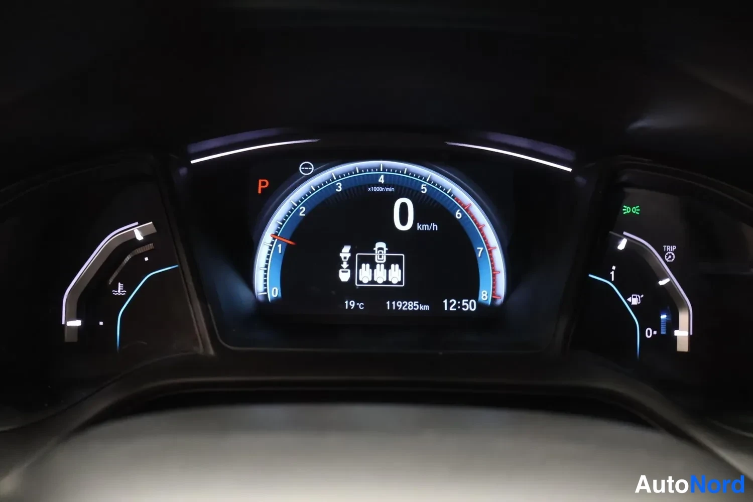 Yksityiskohta: Kojelaudan digitalinen tietoneste: nopeus 0 km/h, vaihde P, lukema 119285 km, ulkolämpötila 19 °c ja aika 12:50.