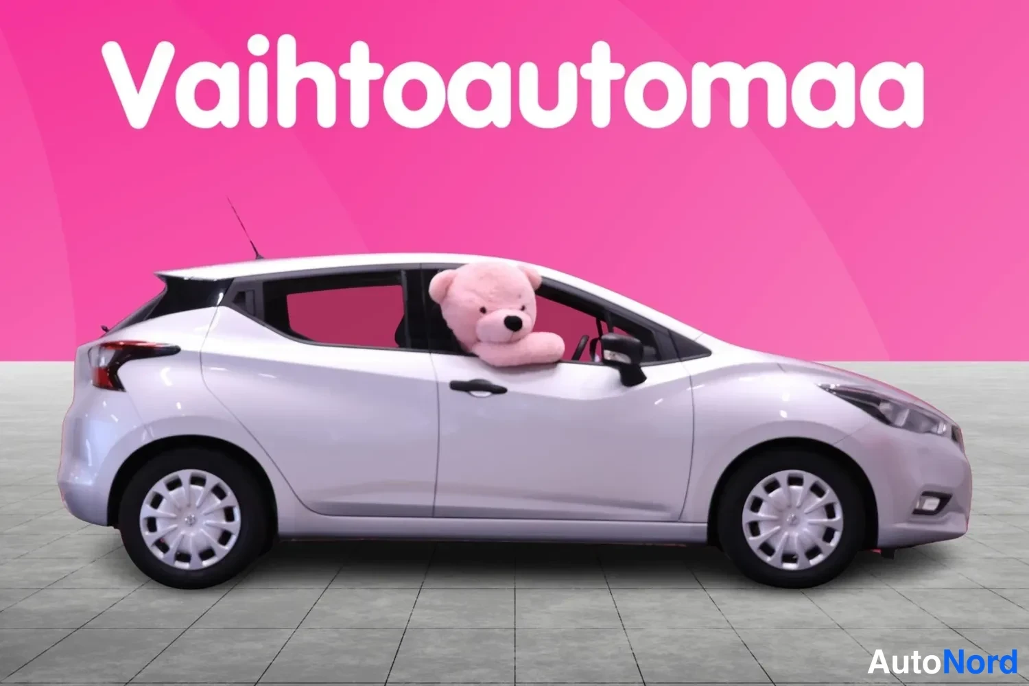 Etuviisto, nokka oikealle: Valkoinen Nissan Micra etukulmasta, keula oikealla, nallekeippo ikkunassa pinkissä studioympäristössä