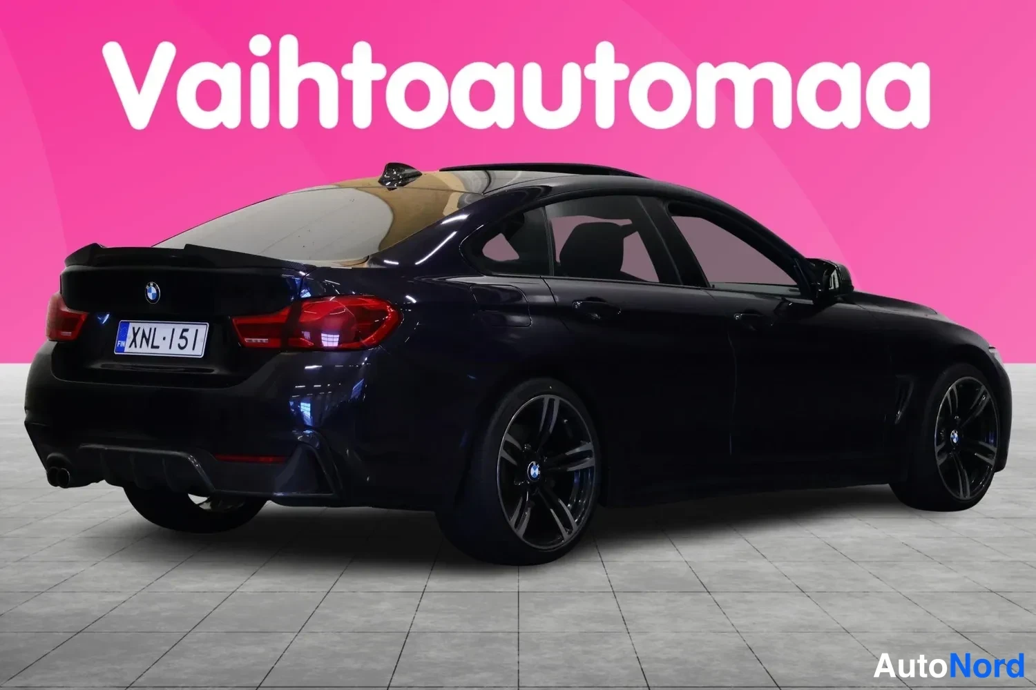 Takaviisto: Auton takakulma: tummansininen BMW, rekisteritunnus, näkyy oikealta takaa pinkkiaustaista taustaa vasten.