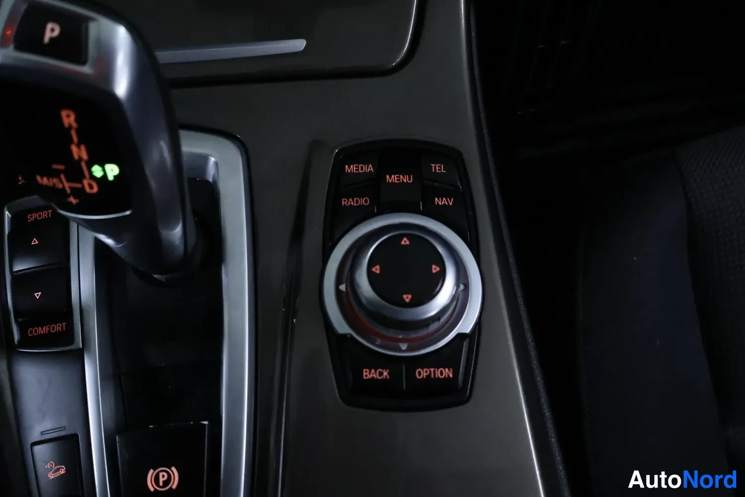 Muu: Keskikonsolin lähikuva: automaattivaihteen valitsin ja iDrive-ohjainknob painikkeineen (MEDIA, RADIO, MENU, TEL, NAV, BACK, OPTION).
