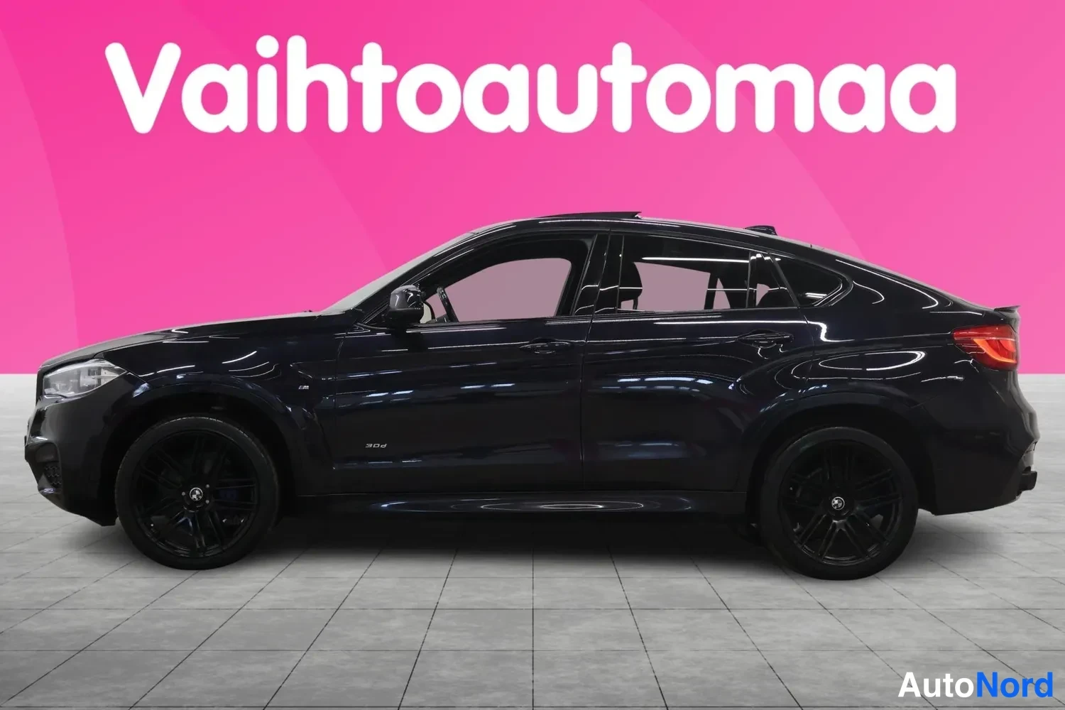 Etuviisto, nokka vasemmalle: Musta BMW X6:n kylki etuosasta näkyvällä keulalla, mustat vanteet, pinkki tausta.