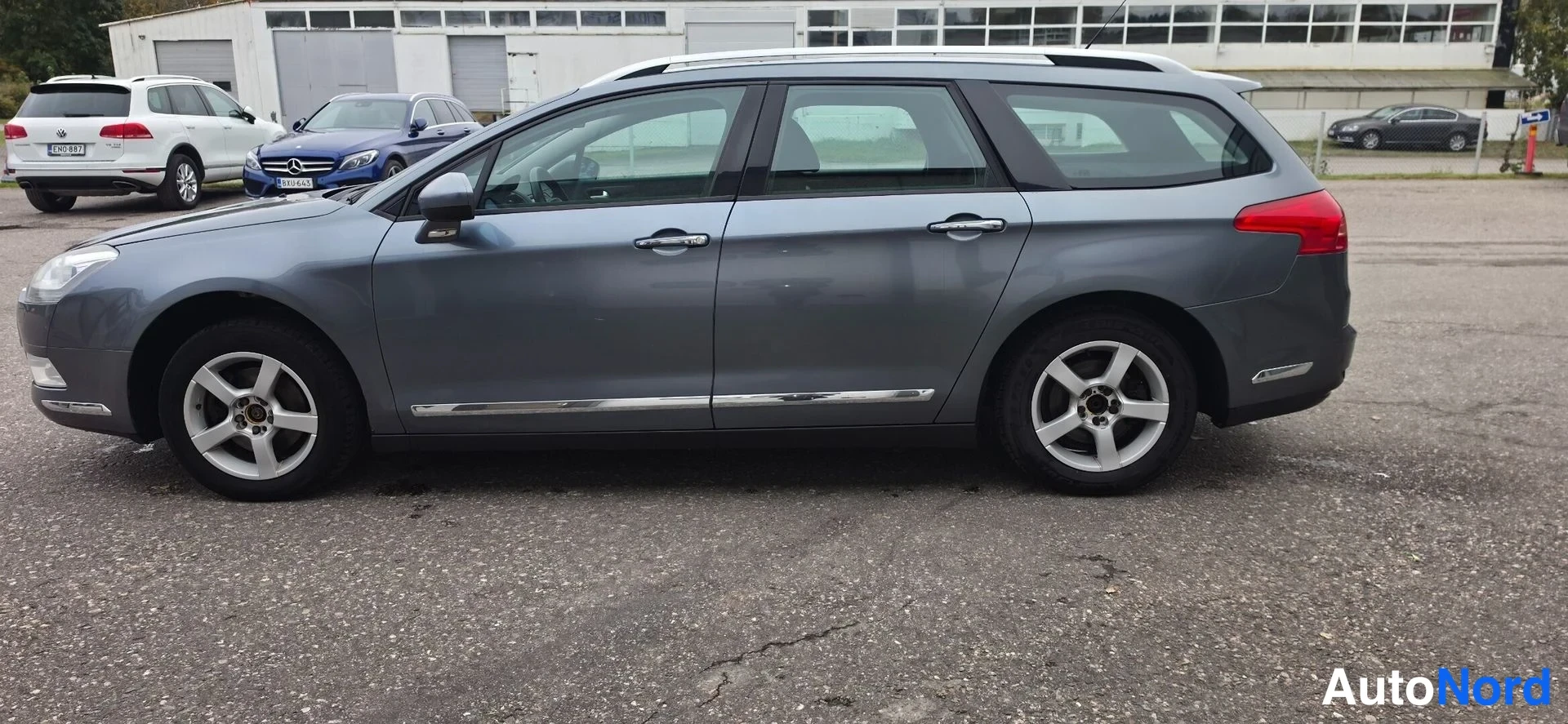 Etuviisto, nokka vasemmalle: Auton etukulma vasemmalta: harmaa farmariauto, jossa kromilistat ovissa.