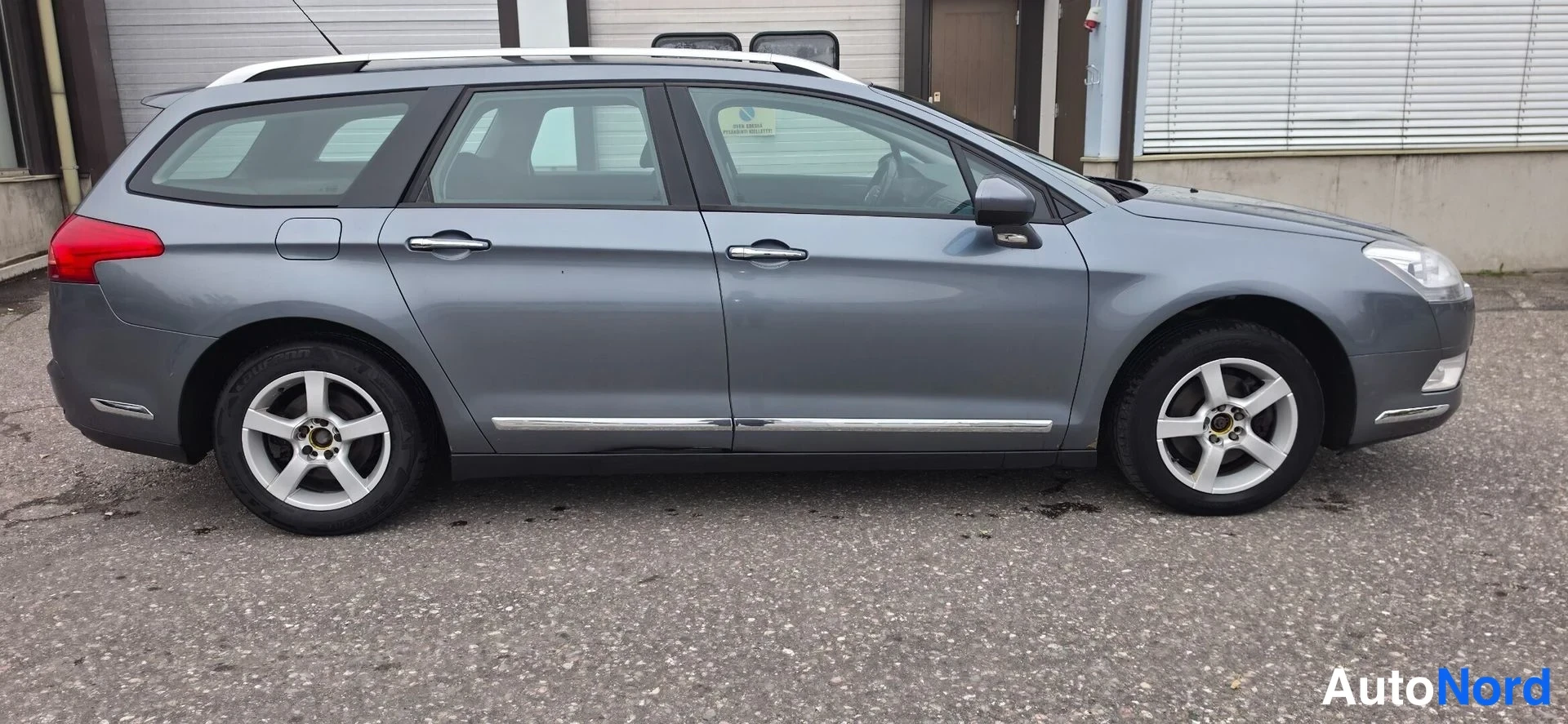 Etuviisto, nokka oikealle: Auton etukulma oikealta: harmaa farmariauto, kattoon asennetut kaiteet ja kromilistat ovissa.