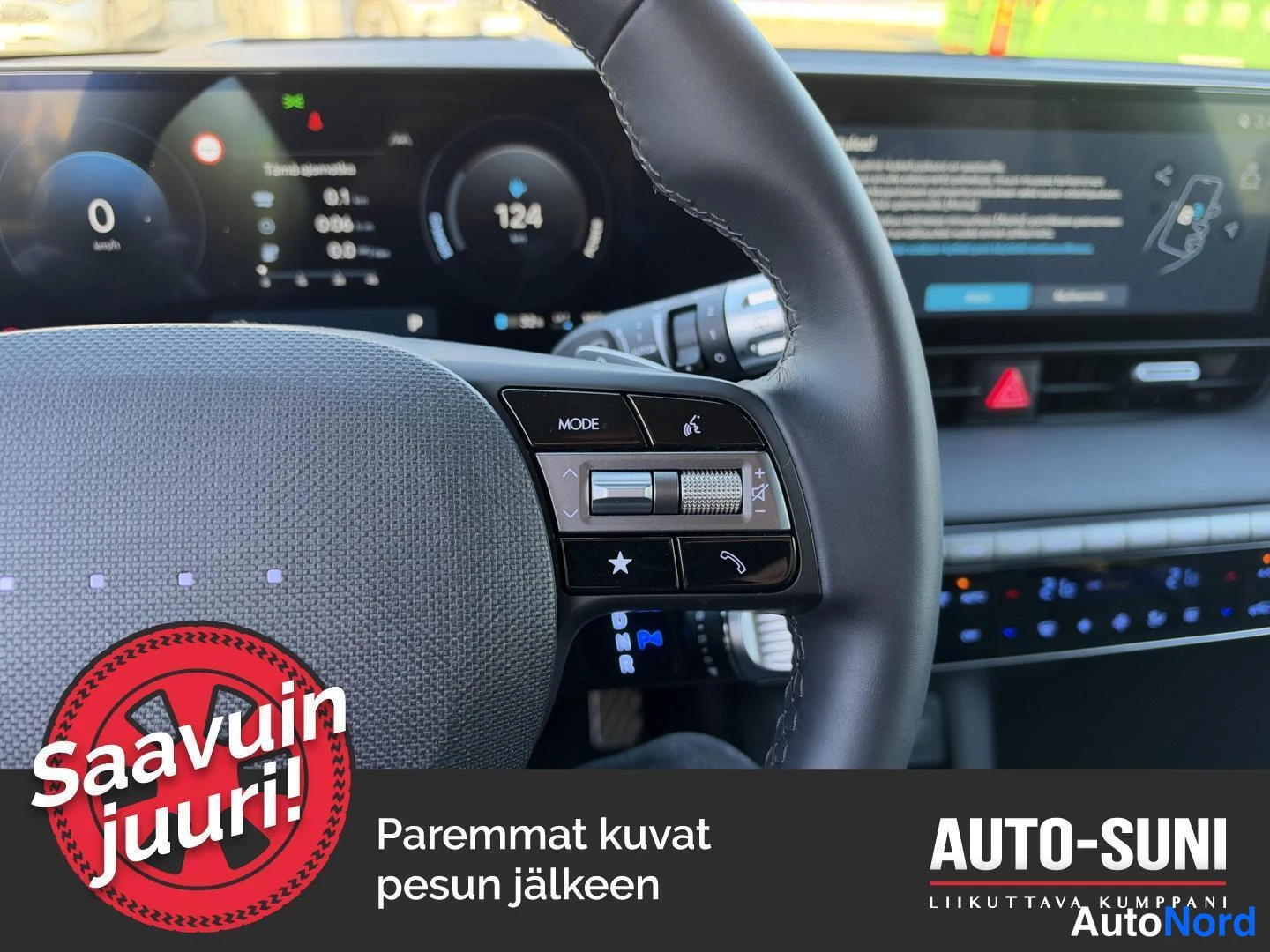 Muu: Hyundai IONIQ 5:n ohjauspyörän oikean puolen MODE-painike ja ääniohjausnappi, taustalla infotainment-näyttö ja ilmastointipaneeli näkyvissä.