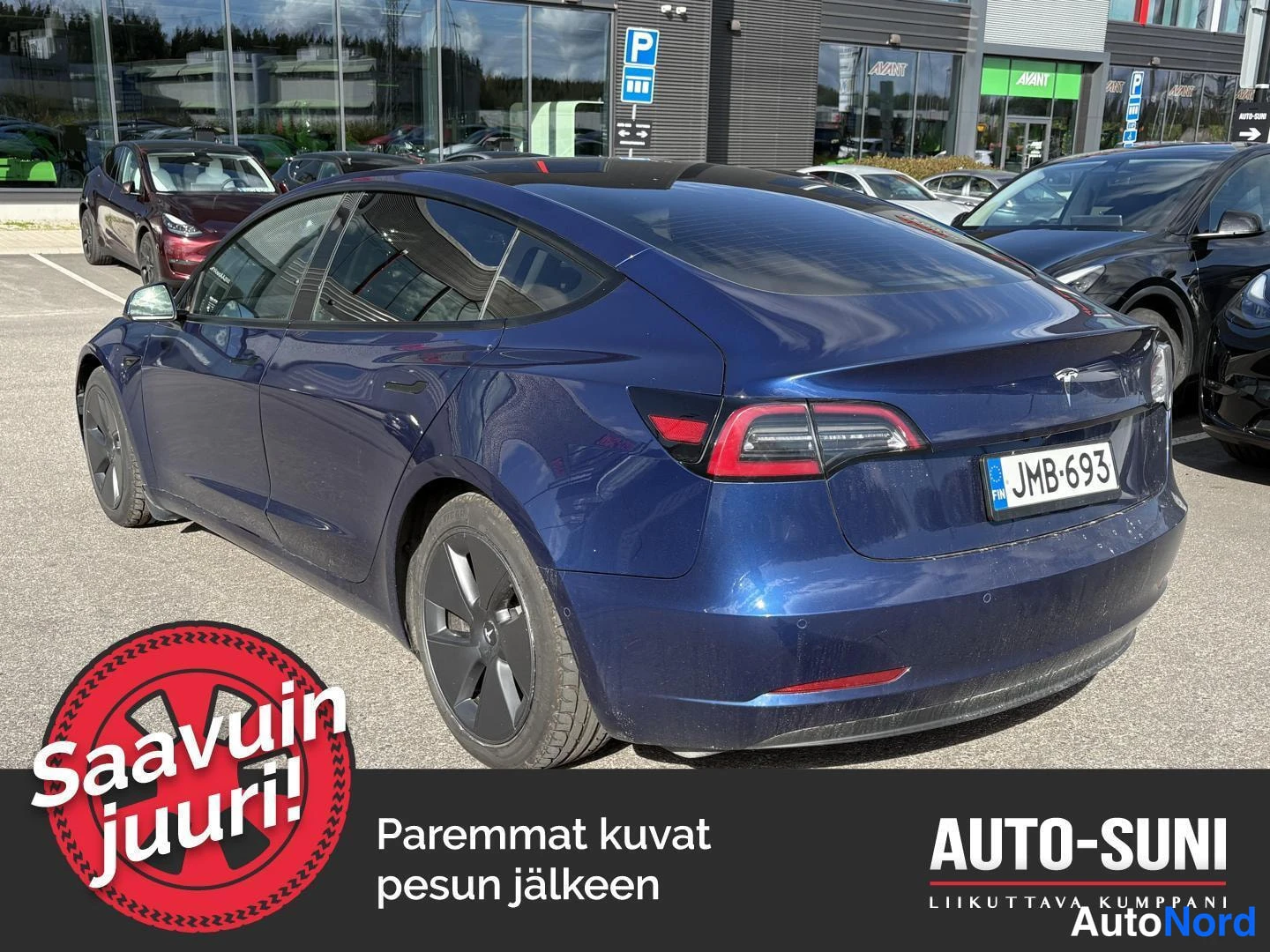 Takaviisto: Tummansininen Tesla Model 3 takaviistöstä oikealta, rekisteri, parkkihallilla muiden autojen seassa.