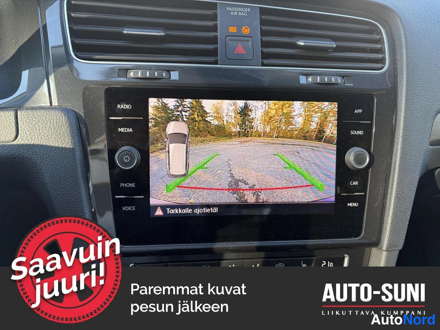 Yksityiskohta: Kojelaudan infotainment-näyttö käänteiskuvanäkymässä: auton kuva ja parkointituet, tekstinä 'Tarkkaile ajotietä!'.