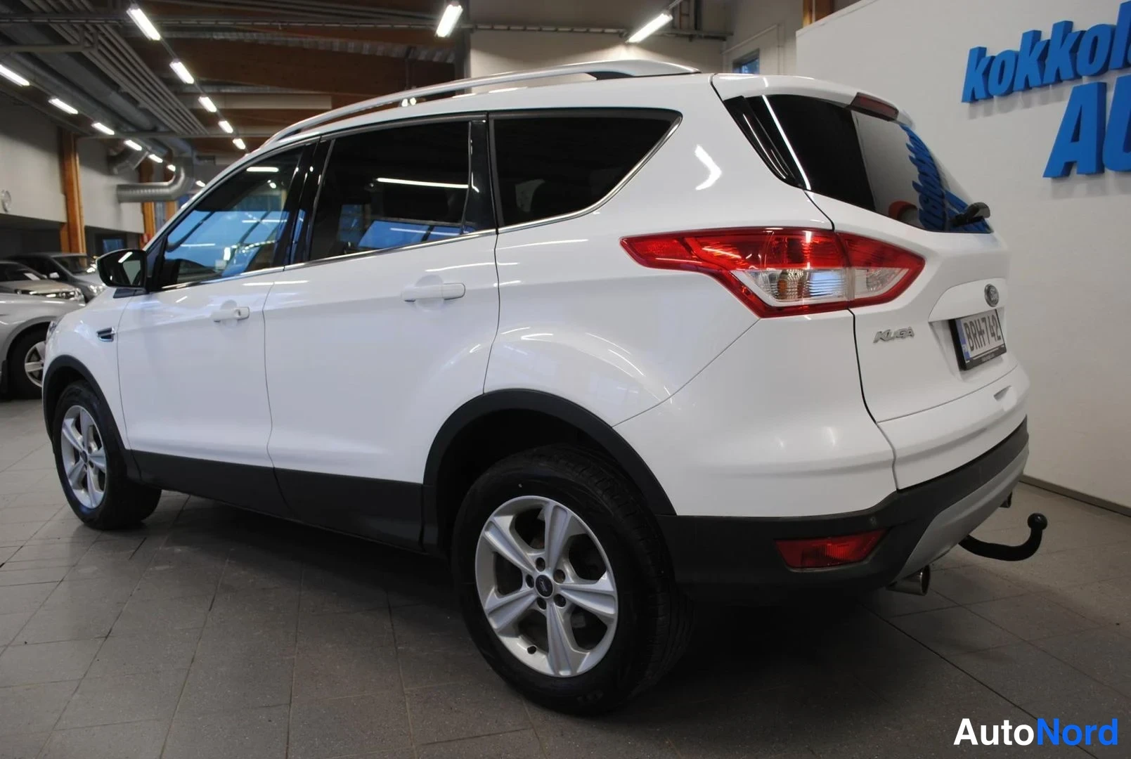 Takaviisto: Valkoinen Ford Kuga koko autona kylki ja takaosa näkyvissä showroomissa, kattotangot, rekisterikilpi.
