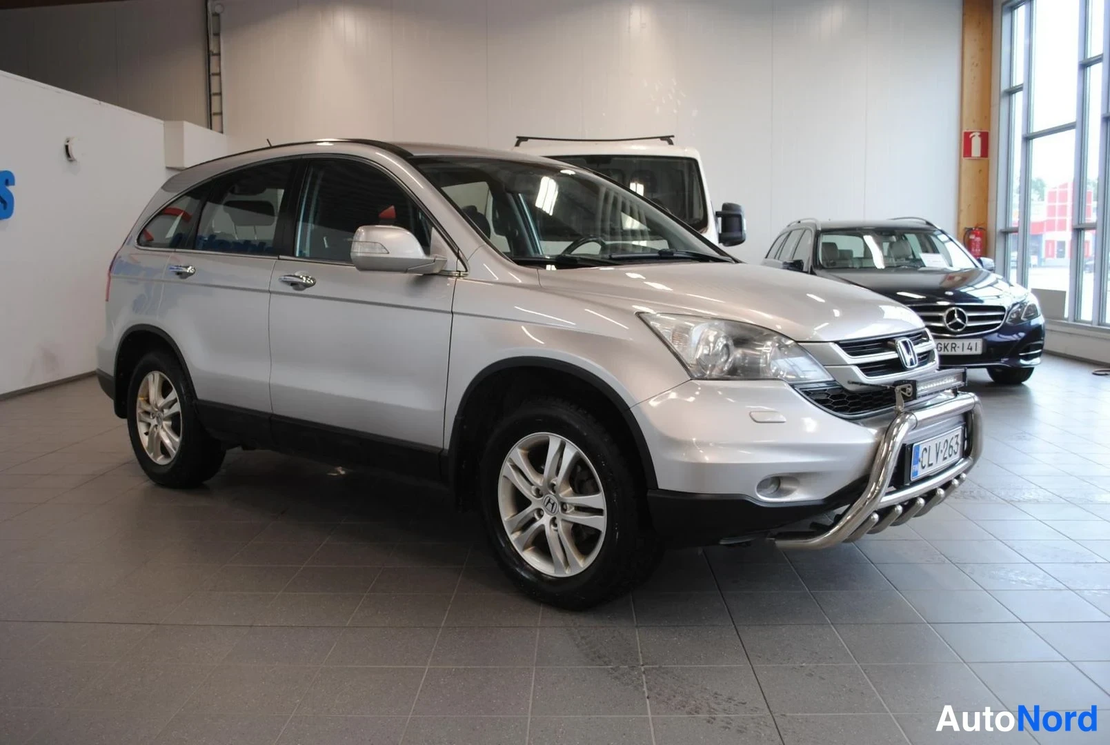 Etuviisto, nokka oikealle: Hopeanvärinen Honda CR-V kuljettajan puolen takaa kolmesta neljäsosasta kuvattuna autosalissa; keulakaari ja rekisterikilpi näkyvät.