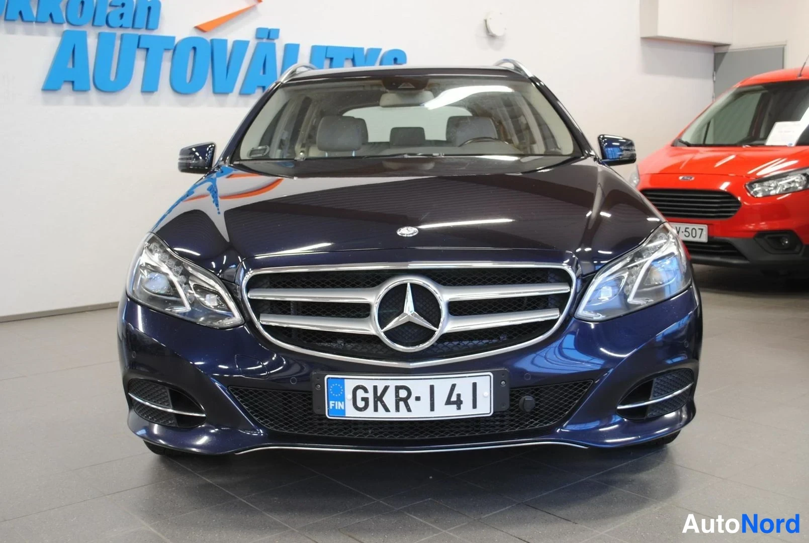 Edestä: Tummansininen Mercedes-Benz suoraan edestä kuvattuna, jäähdytinsäleikkö ja Mercedes-tähti keskellä, rekisteritunnus.