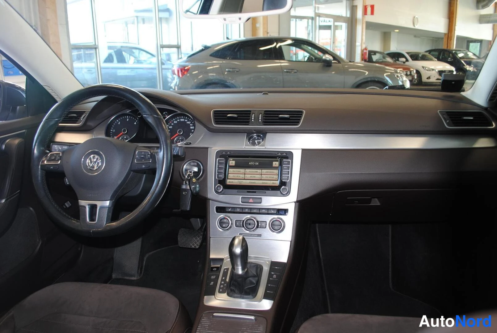 Muu: Ohjaamon näkymä eteenpäin: Volkswagen-ohjauspyörä, mittarit ja keskiössä radionäyttö sekä automaattivalitsin, ikkunoista varikko.
