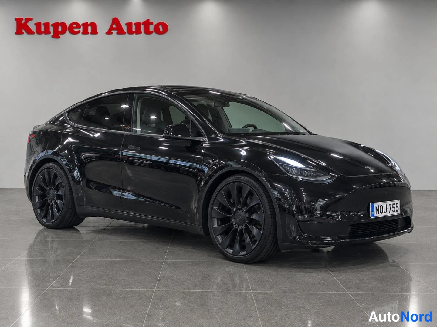 Etuviisto, nokka oikealle: Musta Tesla Model Y etu kolmannesvasen kuvakulmassa autosalissa, rekisterikilpi.