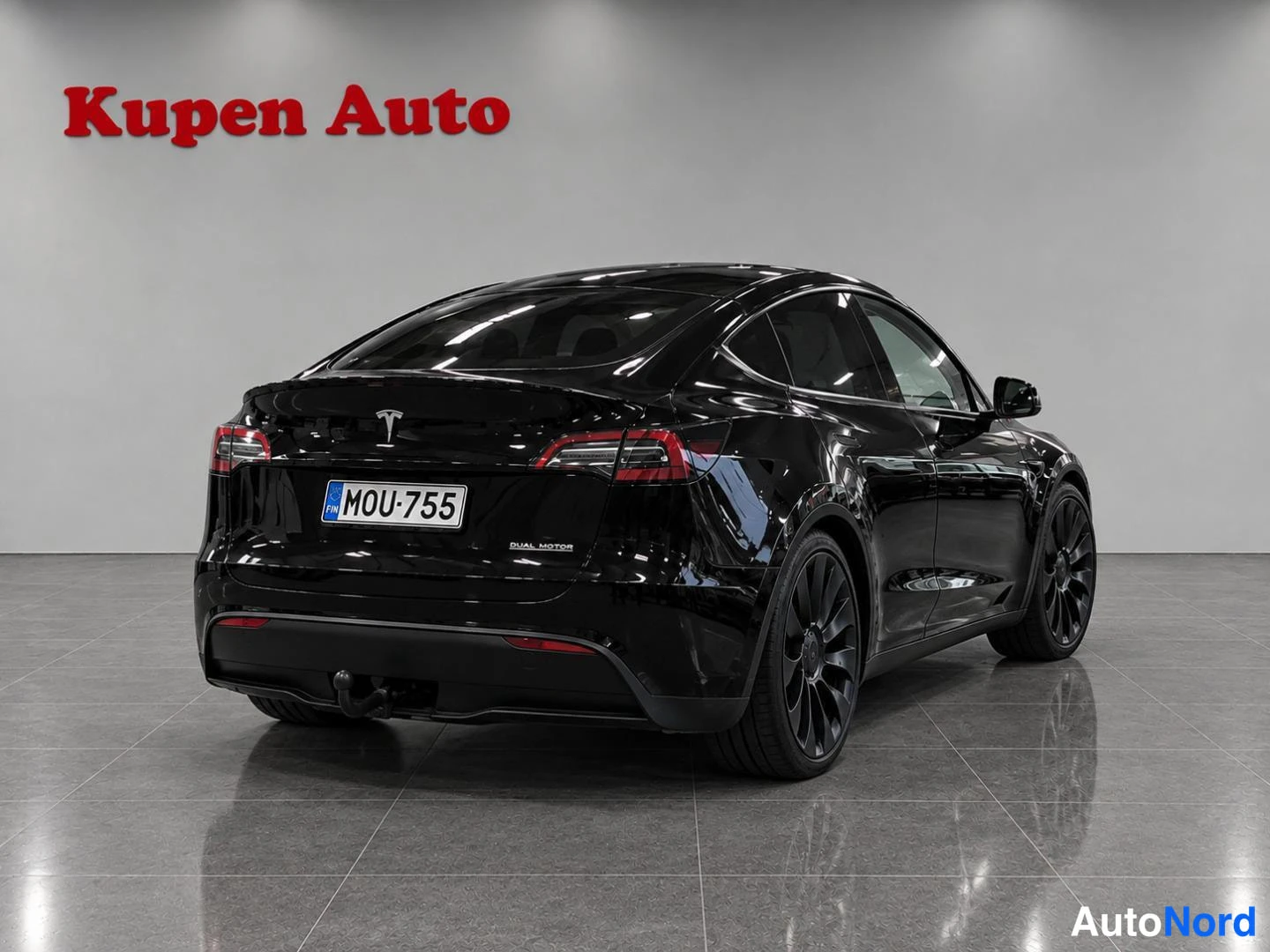 Takaviisto: Musta Tesla Model Y taka kolmannesvasen kuvakulmassa autosalissa, rekisterikilpi.