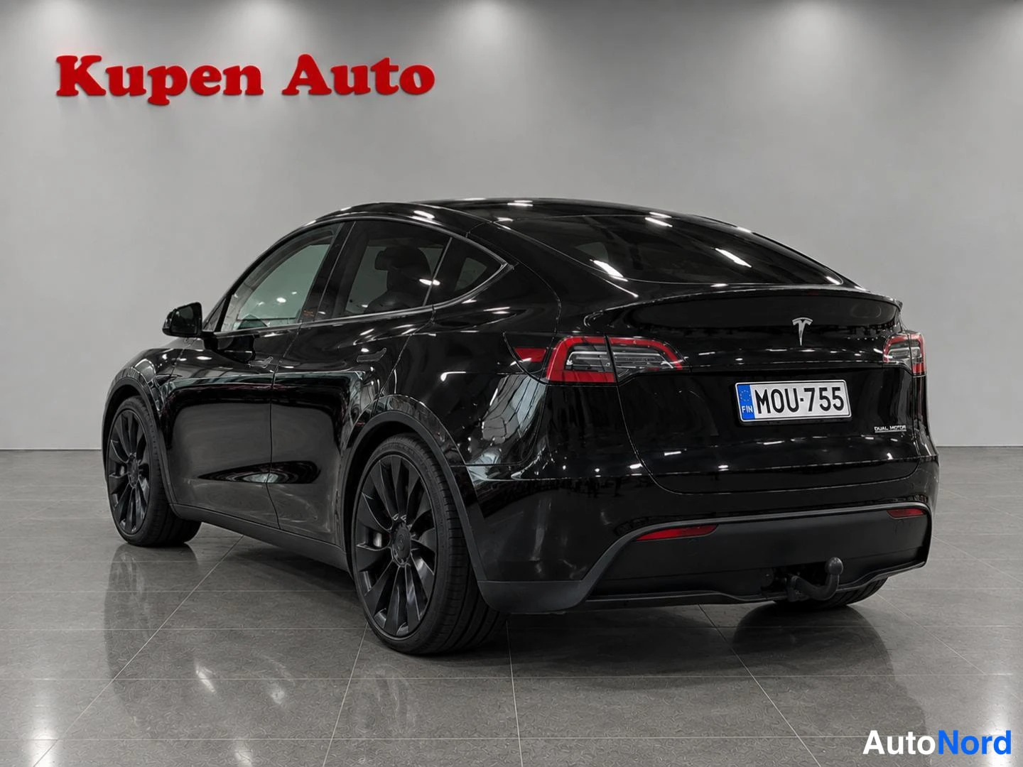 Takaviisto: Musta Tesla Model Y taka kolmannesvasen kuvakulmassa autosalissa, rekisterikilpi.