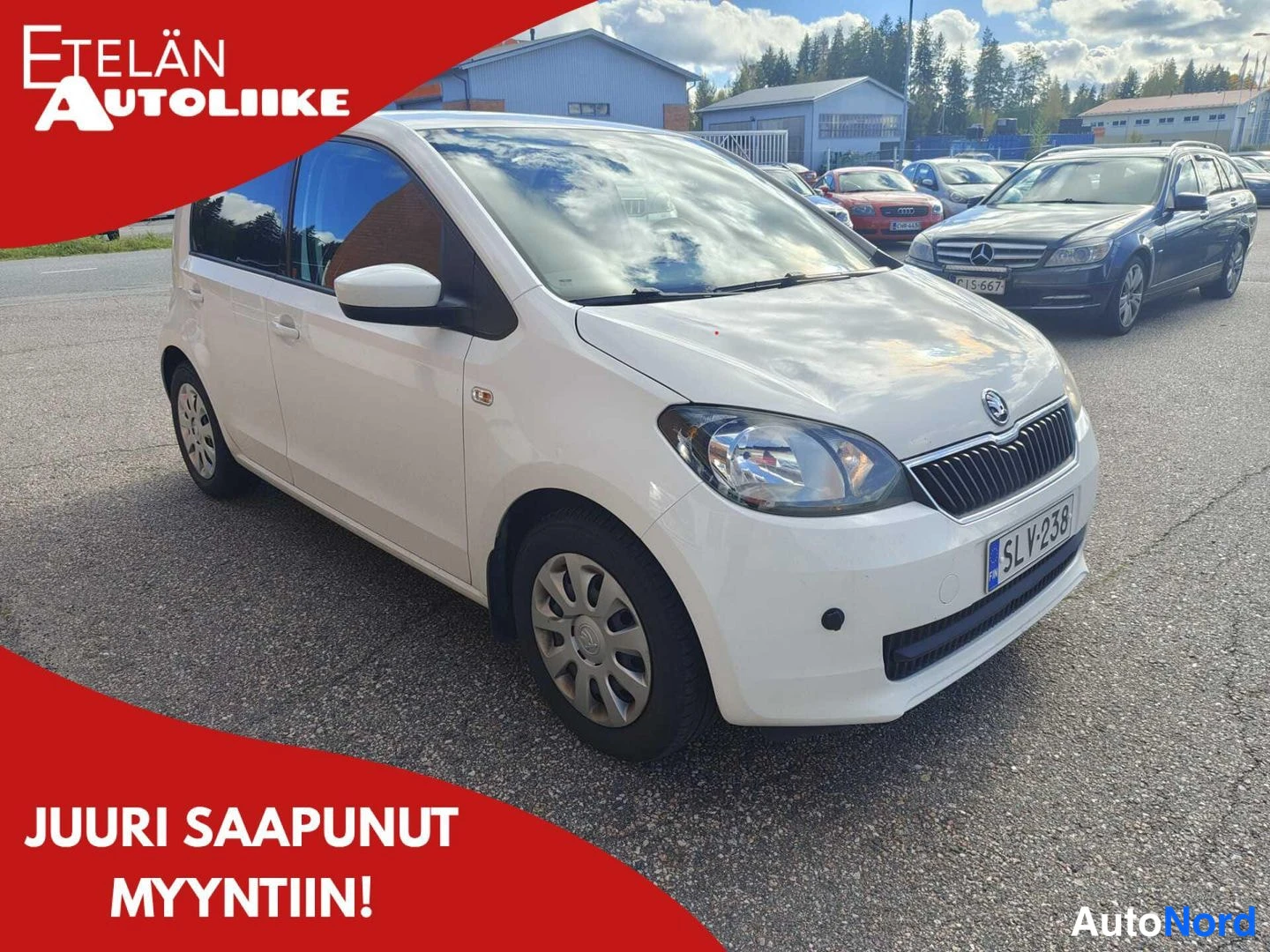 Etuviisto, nokka oikealle: Valkoinen Skoda Citigo esillä autotallin pihalla, kuvattu kolmivirtauksesta keulasta oikealle. Etusäleikkö, valo ja rekisterikilpi näkyvät. Auton koko ko
