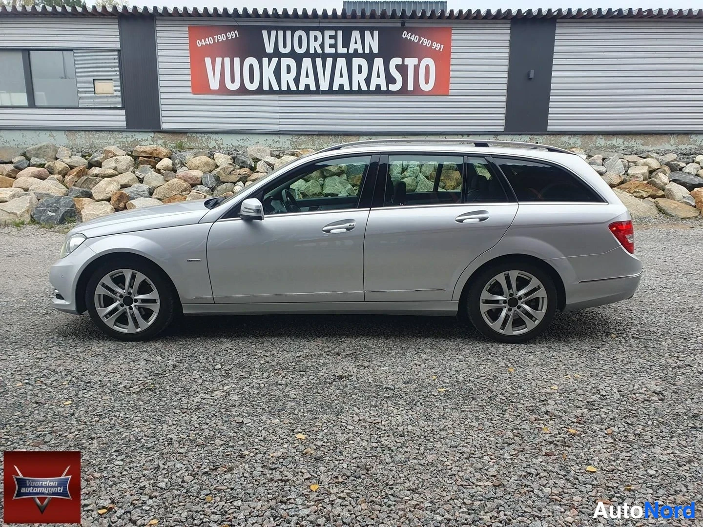 Sivulta: Hopeanharmaa farmariauto sivusta kuvattuna sorapohjaisella paikalla, taustalla kiviaita ja varastorakennus.