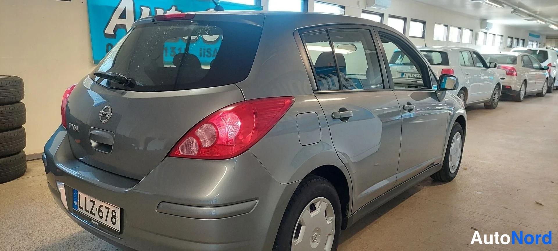 Takaviisto: Hopeanharmaan Nissan Tiidan takapää ja vasen sivu sisätiloissa: takavalot, kannenlukon alue ja -rekisterikilpi näkyvät.